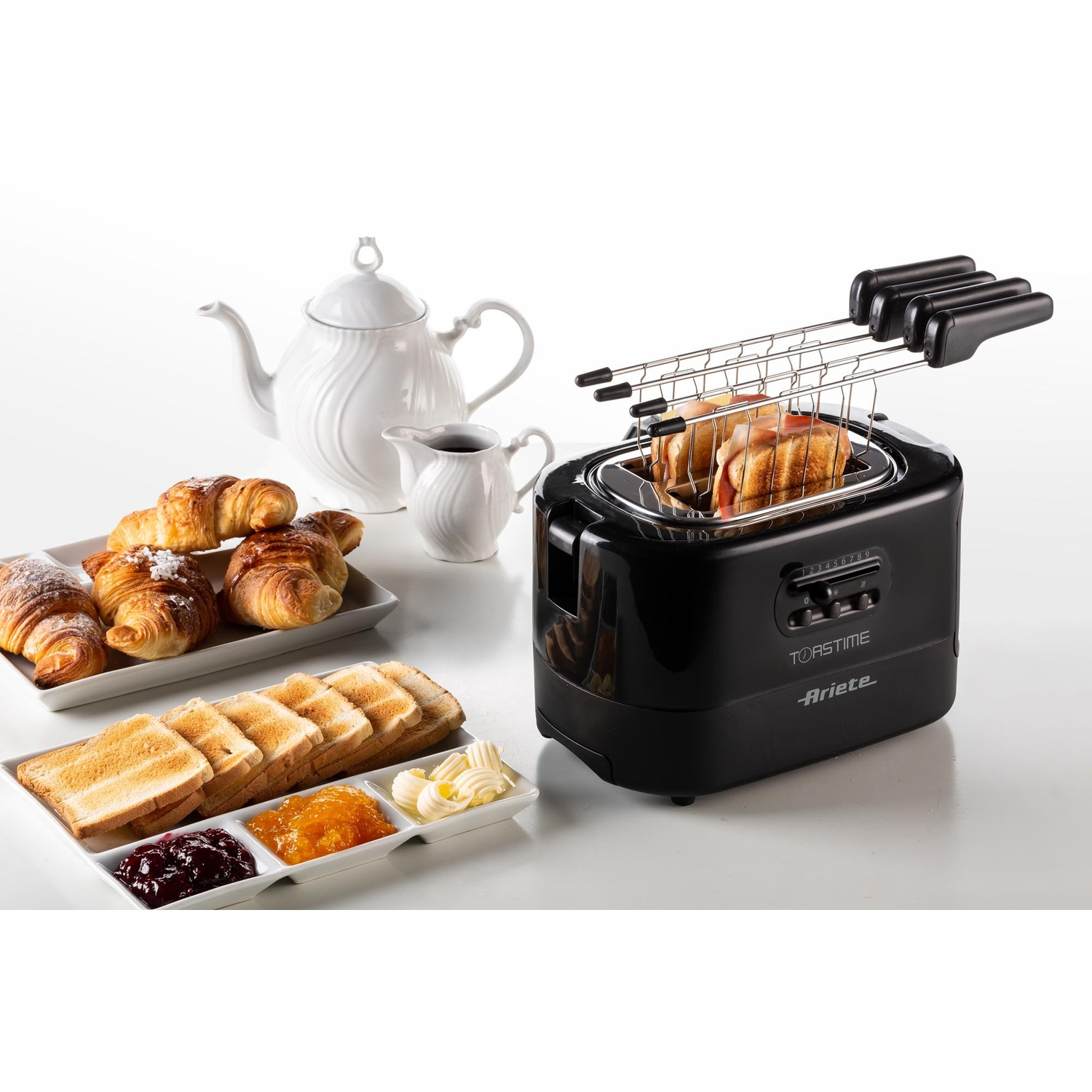 Un tostapane nero con toast, croissant e marmellata su una superficie bianca, con una teiera e un bricco bianchi.