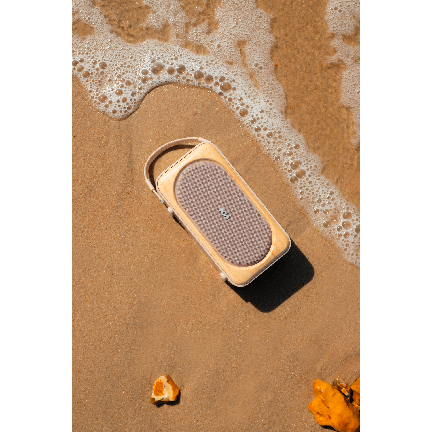 Een luidspreker op een zandstrand met golven. De luidspreker heeft een beige mesh voorkant en een houten frame.