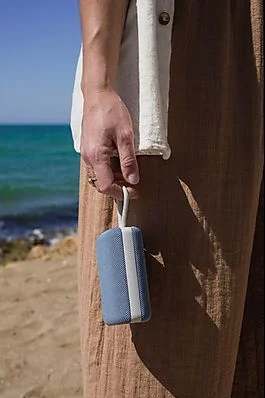 Osoba trzyma mały niebieski głośnik. Tłem jest plaża i ocean.