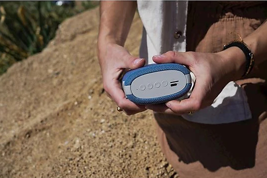 Osoba trzyma niebiesko-szary głośnik przenośny. Tło to piaszczysty teren z roślinami i skałami.