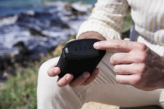 Dłonie trzymają czarny głośnik, dotykając przycisków na górze.