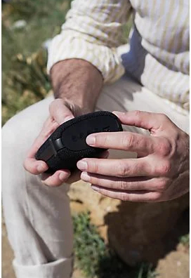 Ręce osoby trzymającej małe, czarne, zaokrąglone urządzenie. Osoba jest na zewnątrz.