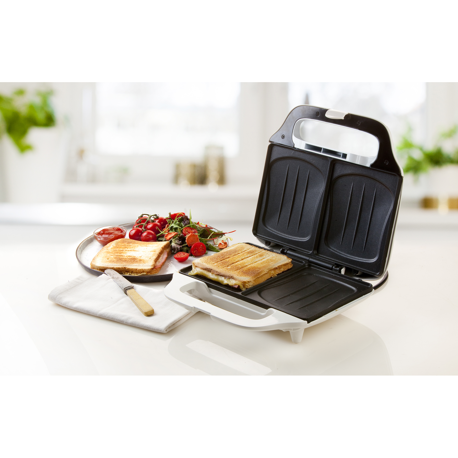 Sandwiches in a sandwich maker, with a plate of food and a knife. White surfaces, bright light.