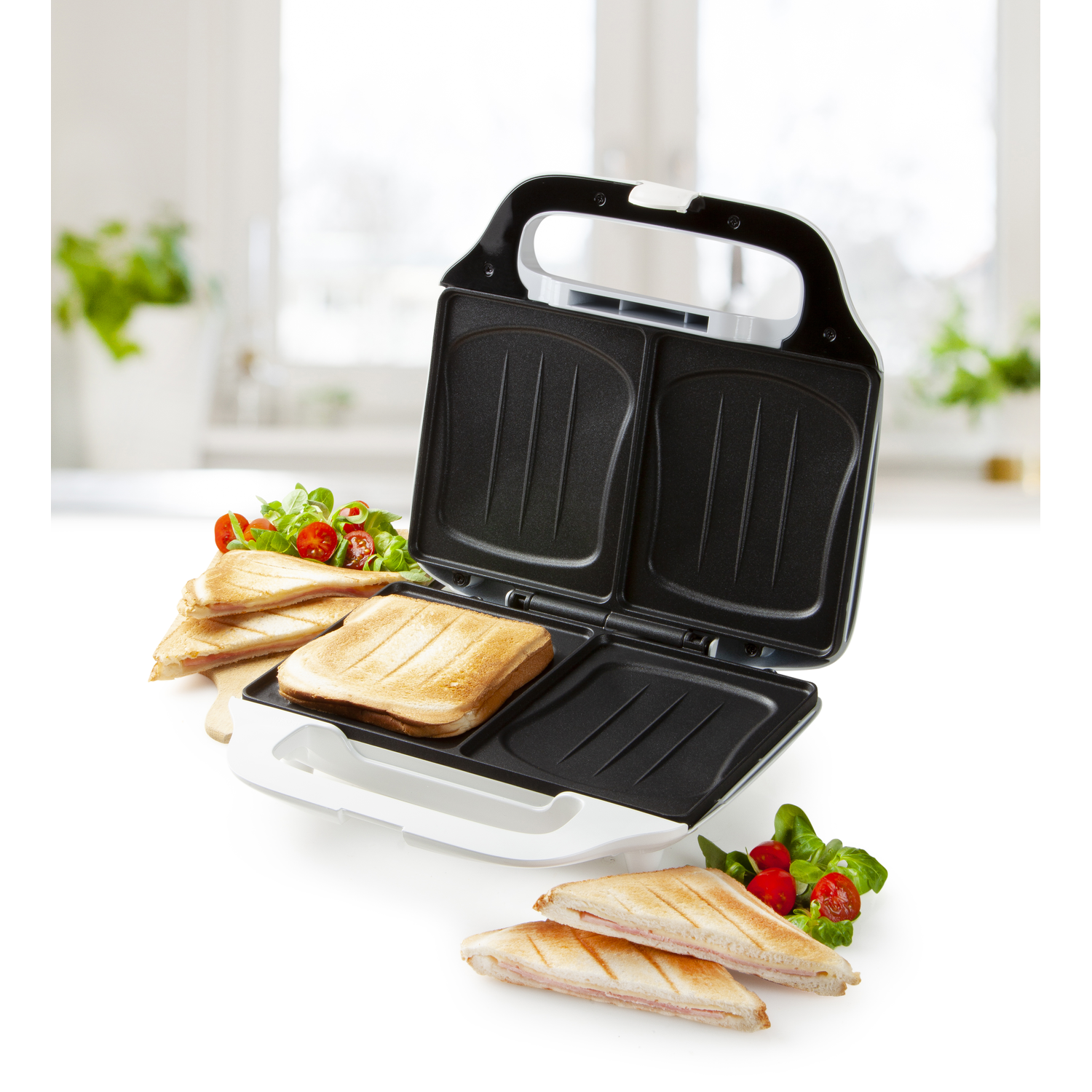 A white and black sandwich maker with sandwiches and garnishes on a white surface.