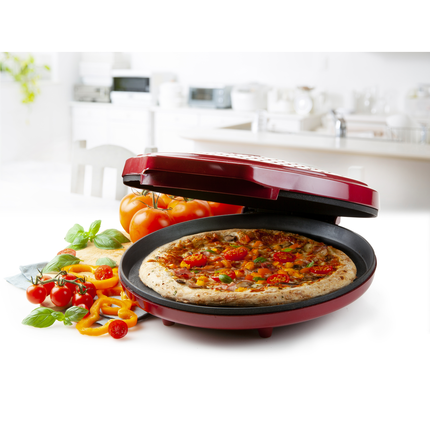 Een pizza in een rode pizzamaker, tomaten, paprika's en basilicum aan de zijkant. Keukenomgeving.