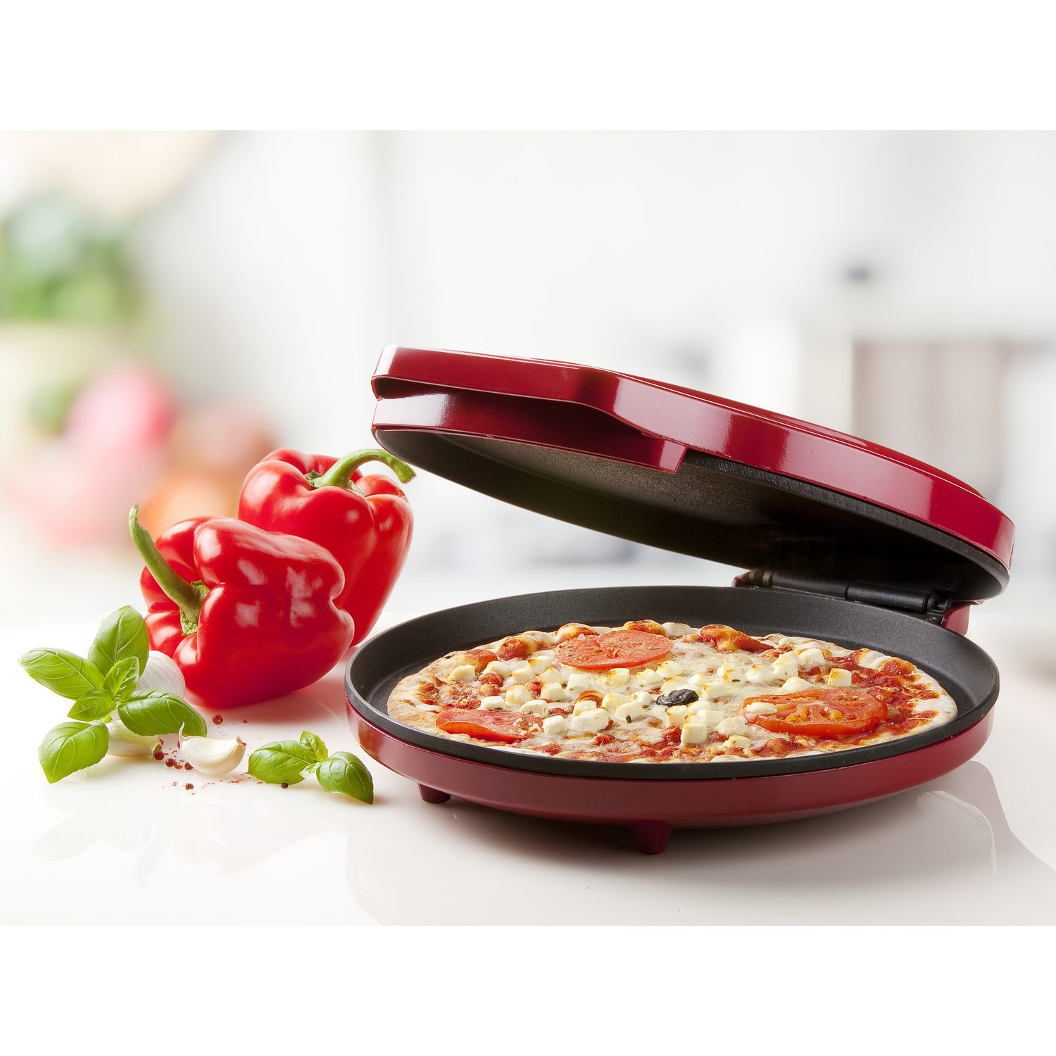 Rode pizzamaker met open deksel, gekookte pizza erin. Rode paprika's en basilicumblaadjes vooraan.