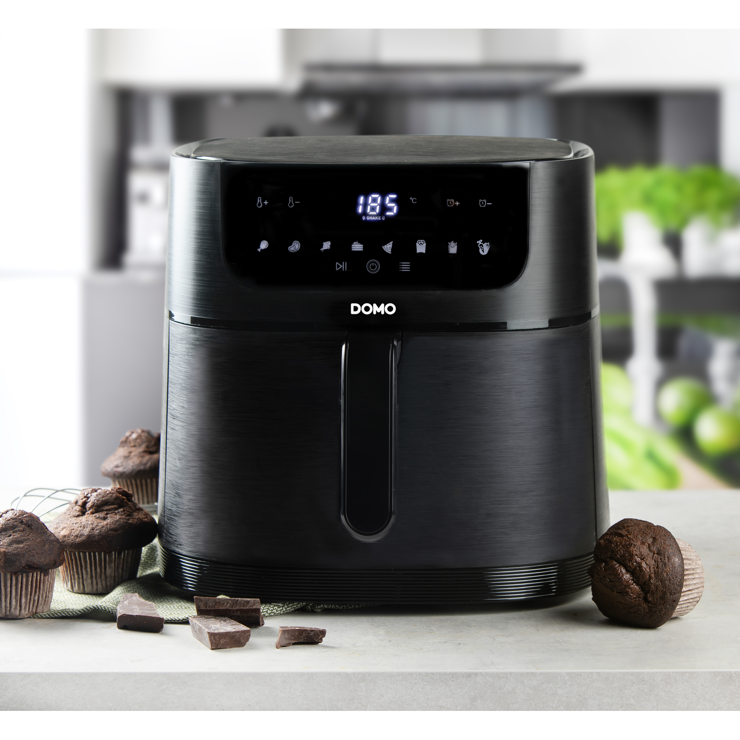 Black air fryer on a counter, with muffins and chocolate pieces nearby.