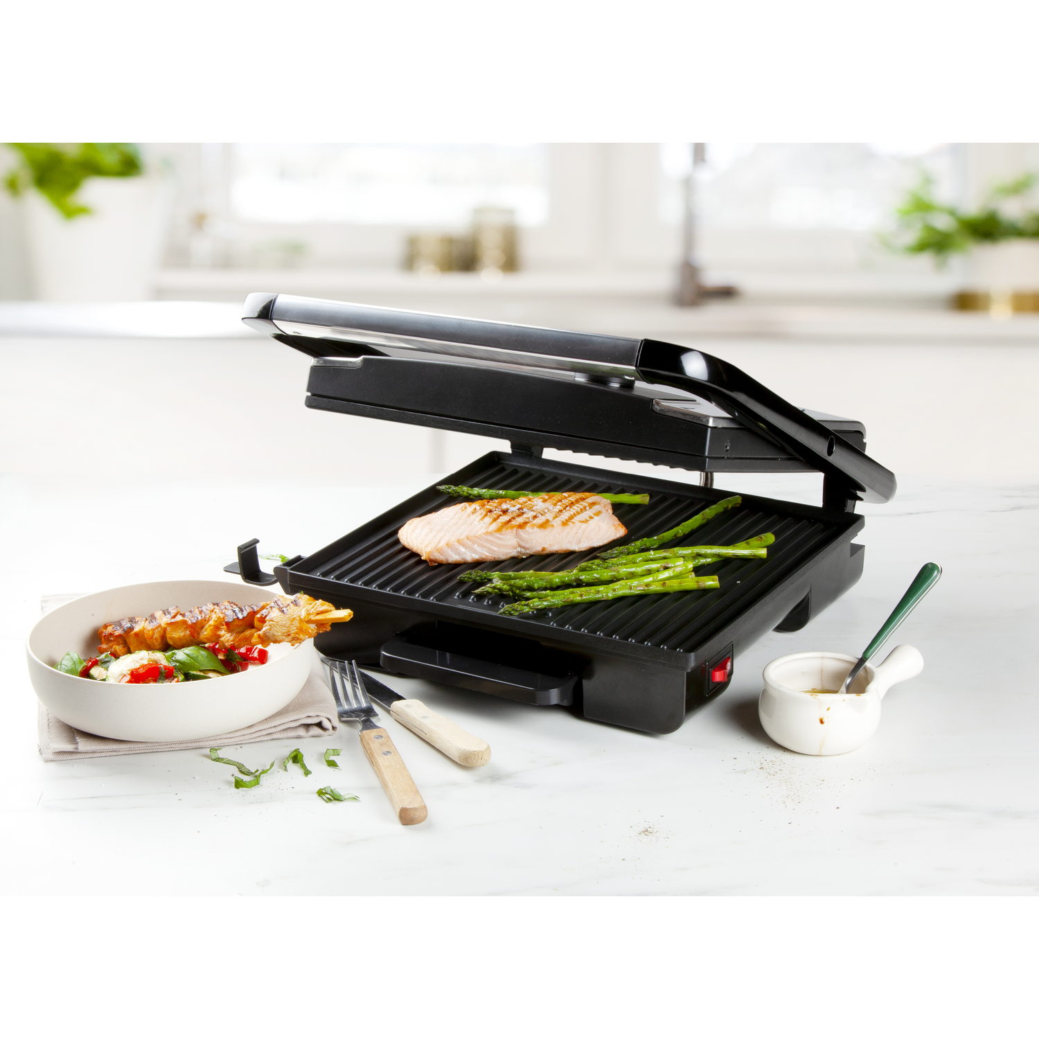 Een grill met zalm en asperges. Een kom met eten, een vork en een mes liggen op tafel. Een sausschaal met een lepel.
