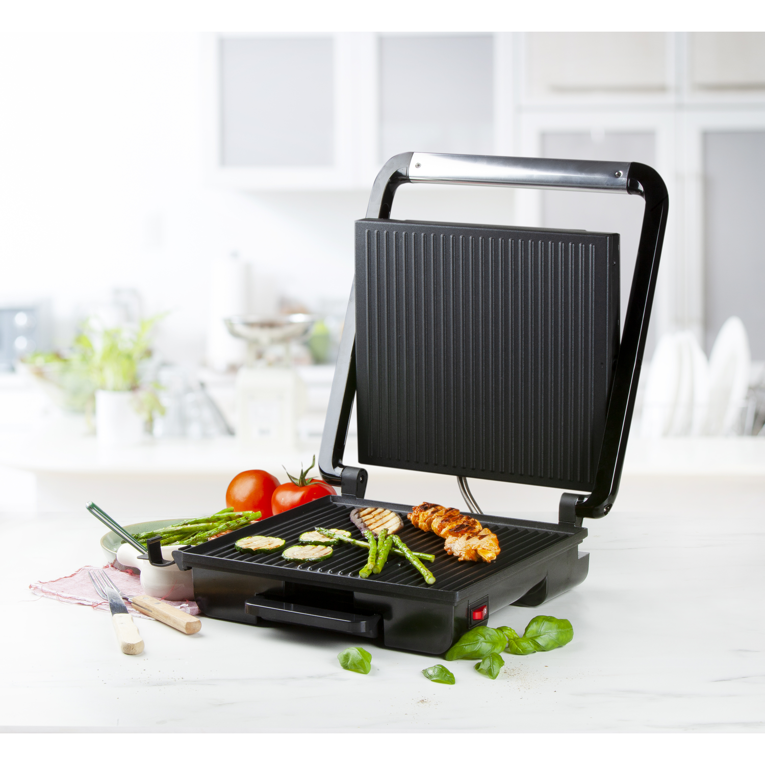 Een zwarte grill met gegrilde groenten en vlees. Tomaten, asperges en basilicum zijn ook aanwezig op een wit oppervlak.