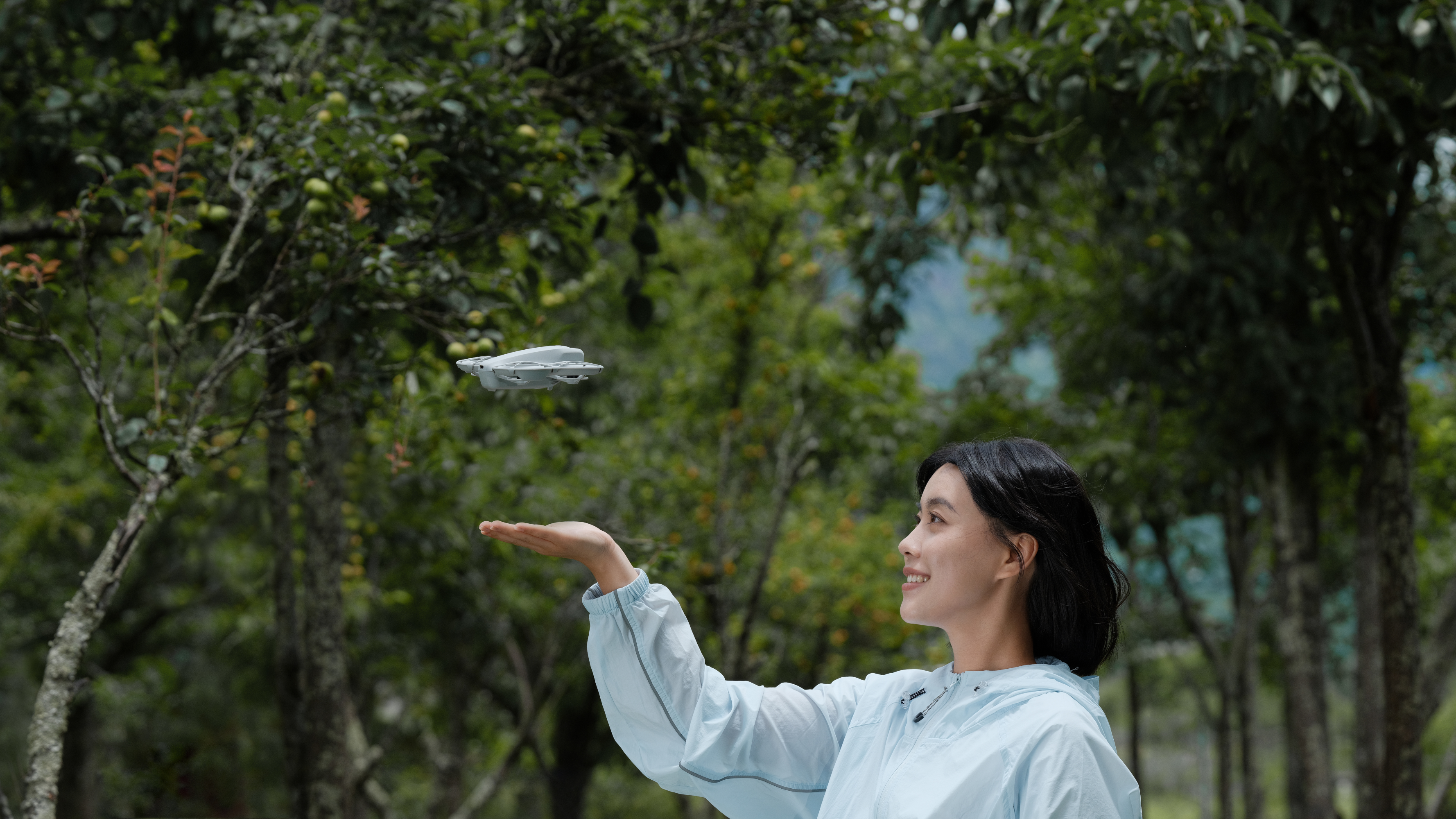 Une femme souriante, regardant un drone planant dans les airs au-dessus de sa main. Des arbres verts en arrière-plan.