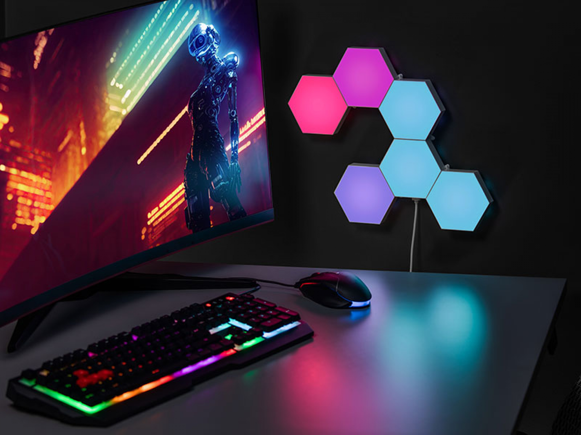 Játékbeállítás egy monitorral, amely egy futurisztikus jelenetet, billentyűzetet és LED-es hatszögletű lámpákat jelenít meg egy sötét felületen.