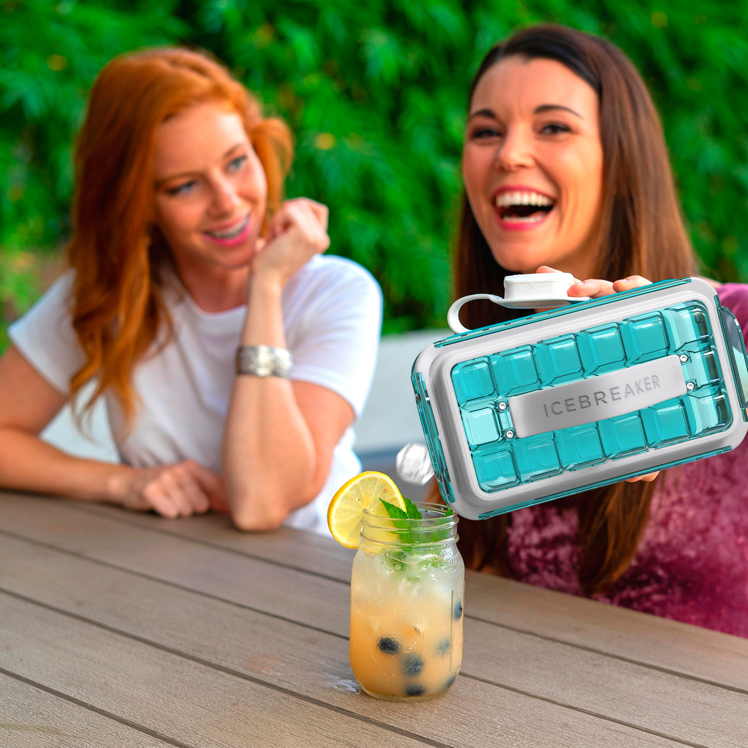 Two women with a cocktail. One holds an ice tray and smiles, the other laughs and looks on.