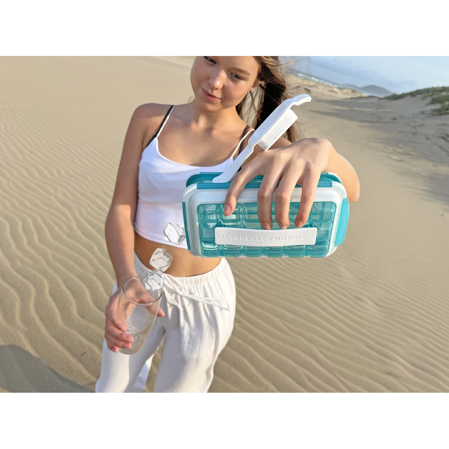 A woman is pouring ice cubes into a glass. She stands on a sandy beach.
