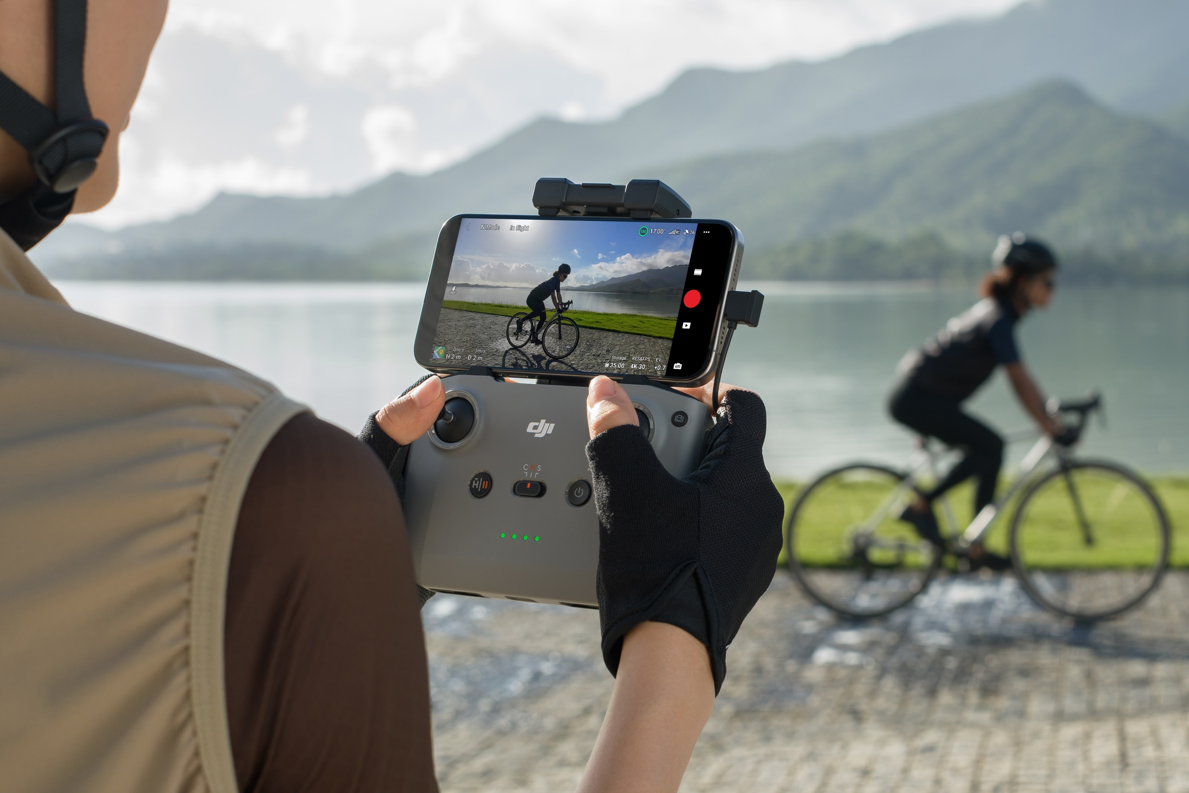 Personne tenant un téléphone montrant un cycliste sur un lac, utilisant une manette DJI. Le fond est la nature.
