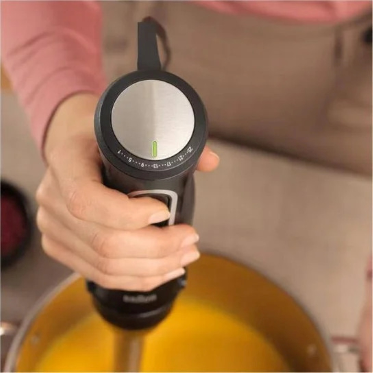 Dłoń trzymająca czarny blender ręczny nad garnkiem z żółtym płynem.