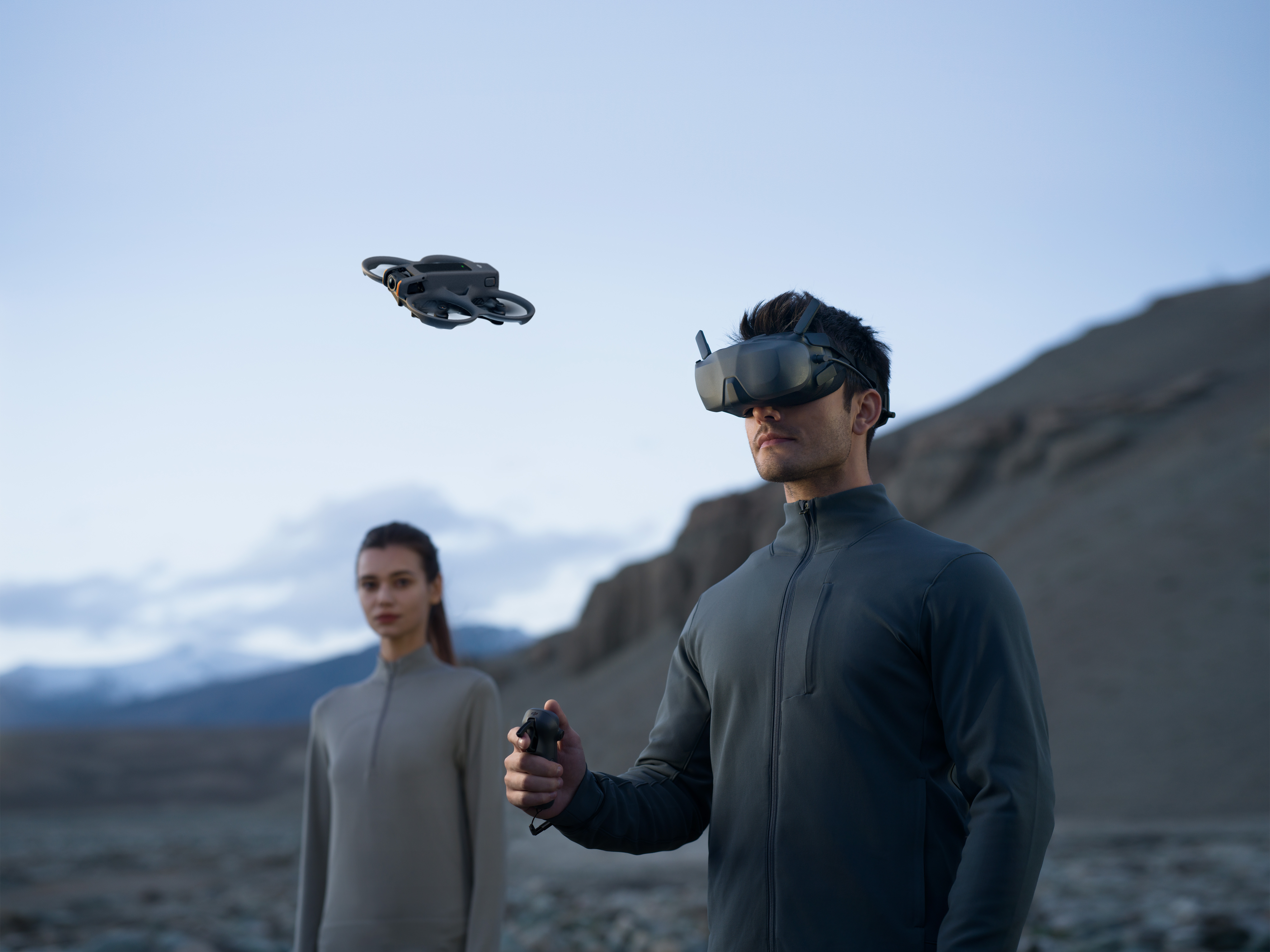 Mann mit VR-Brille, Drohne fliegt. Frau im Hintergrund. Bergiger, bedeckter Hintergrund.