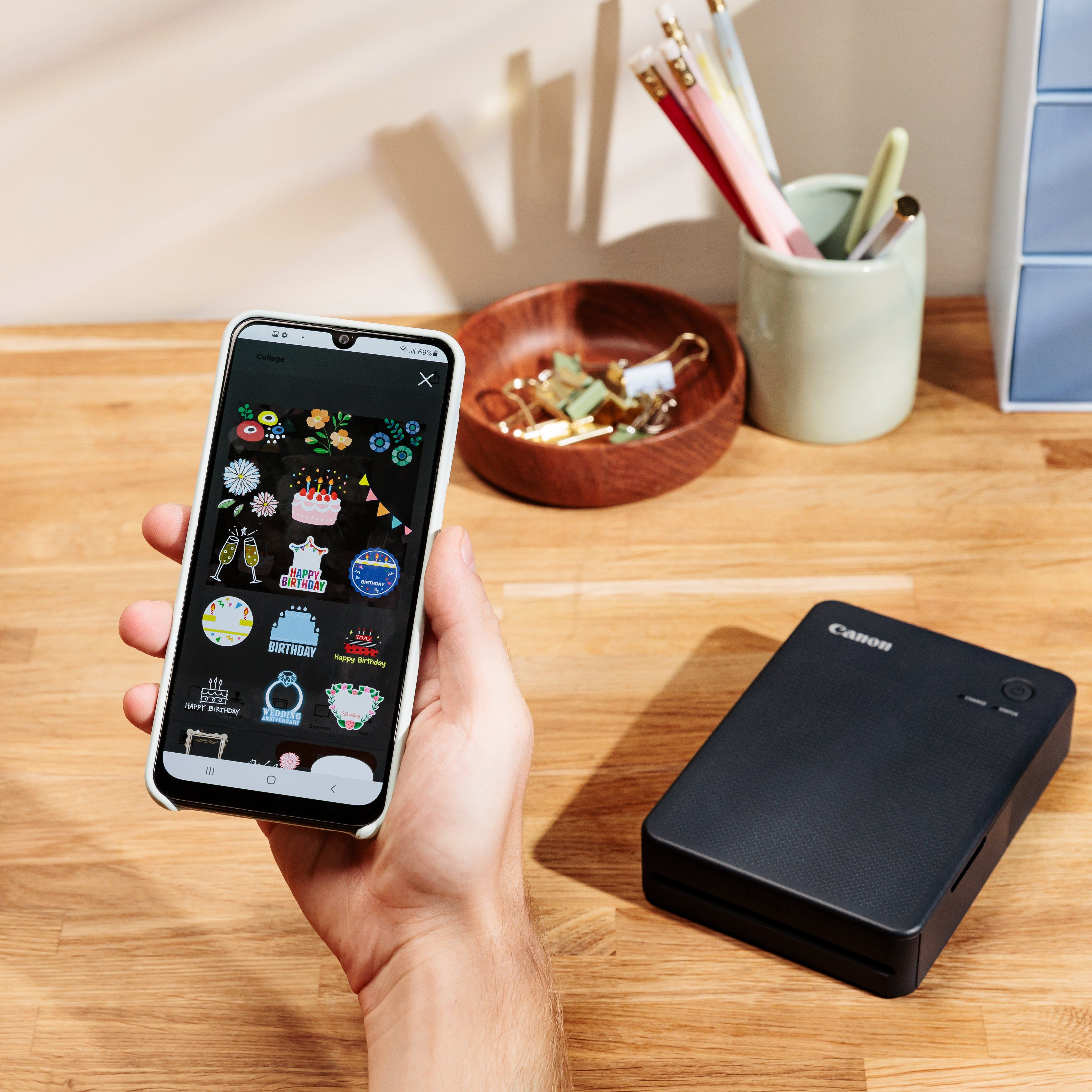 Una mano che tiene uno smartphone che mostra opzioni di adesivi, con una stampante accanto.