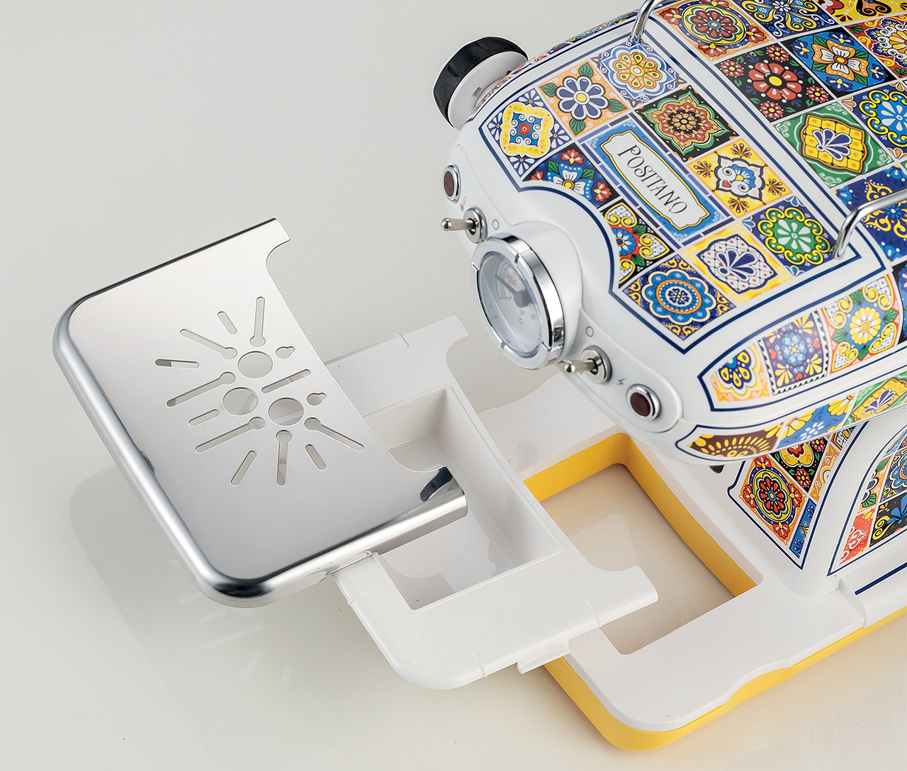 Macchina per caffè espresso con design a piastrelle colorate, vassoio anteriore aperto, superficie bianca.