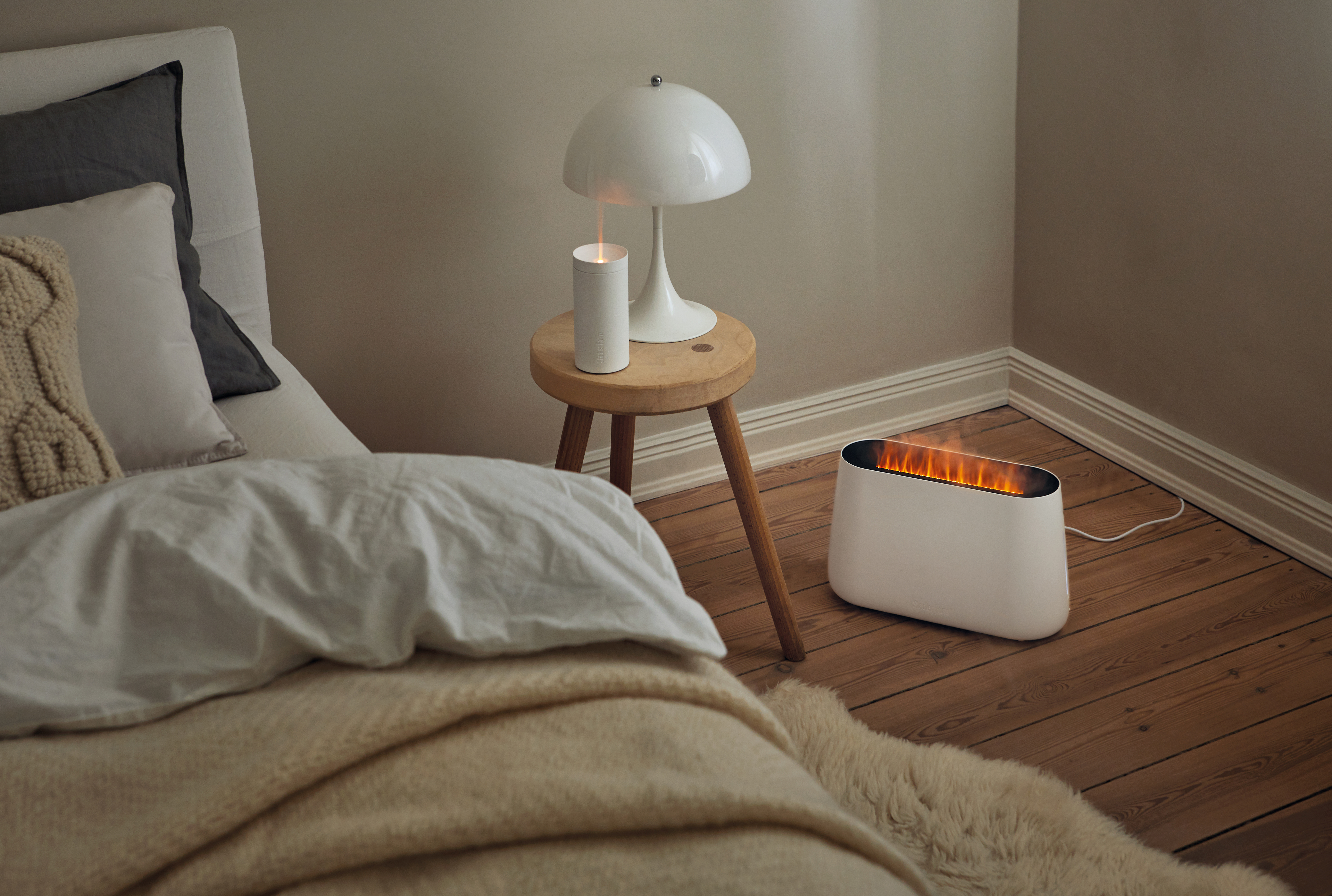 Eine gemütliche Schlafzene mit Lampe, Kerze, kleinem Tisch und einem weißen Kaminofen.