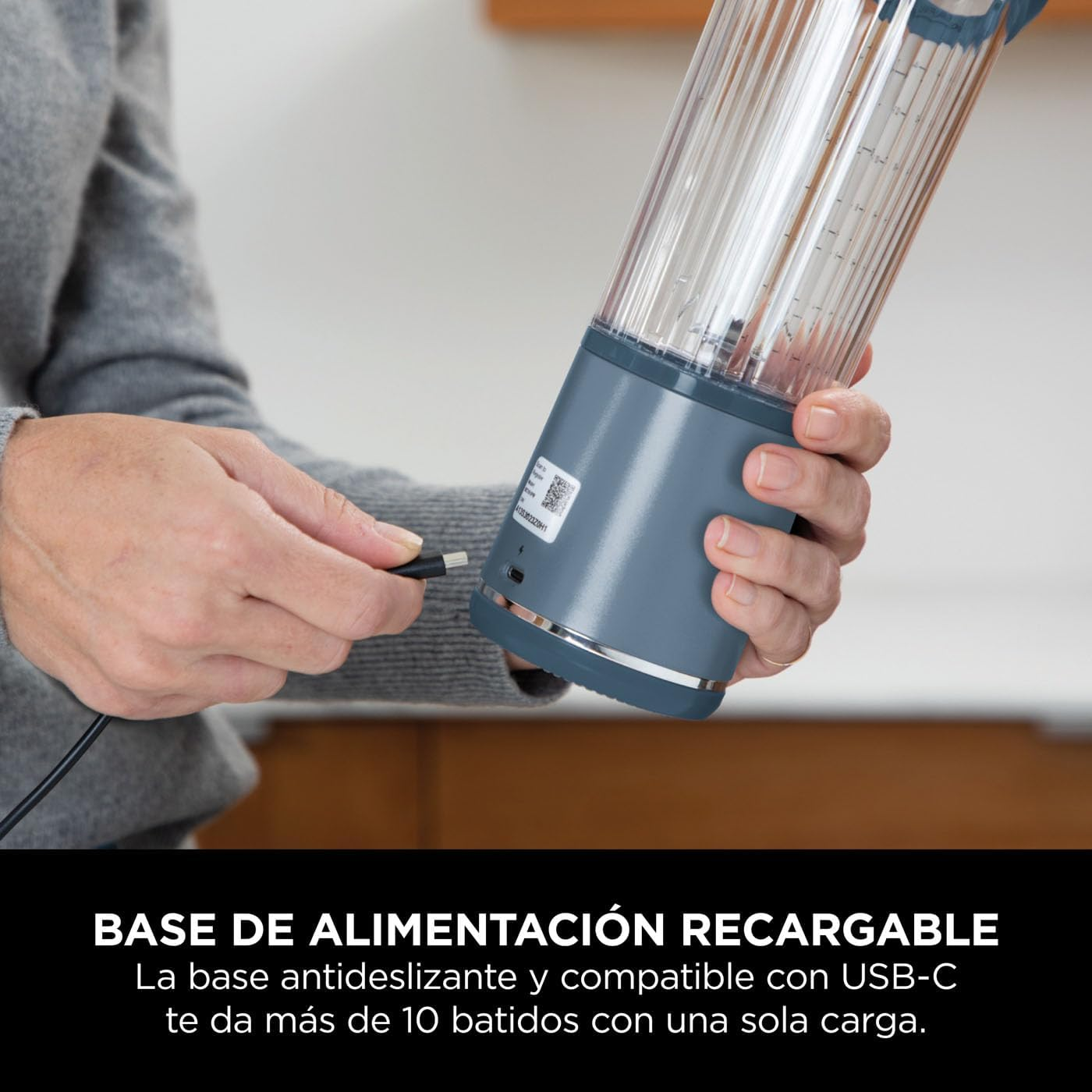 Una mujer sosteniendo una licuadora gris recargable, con un cable de carga conectado.