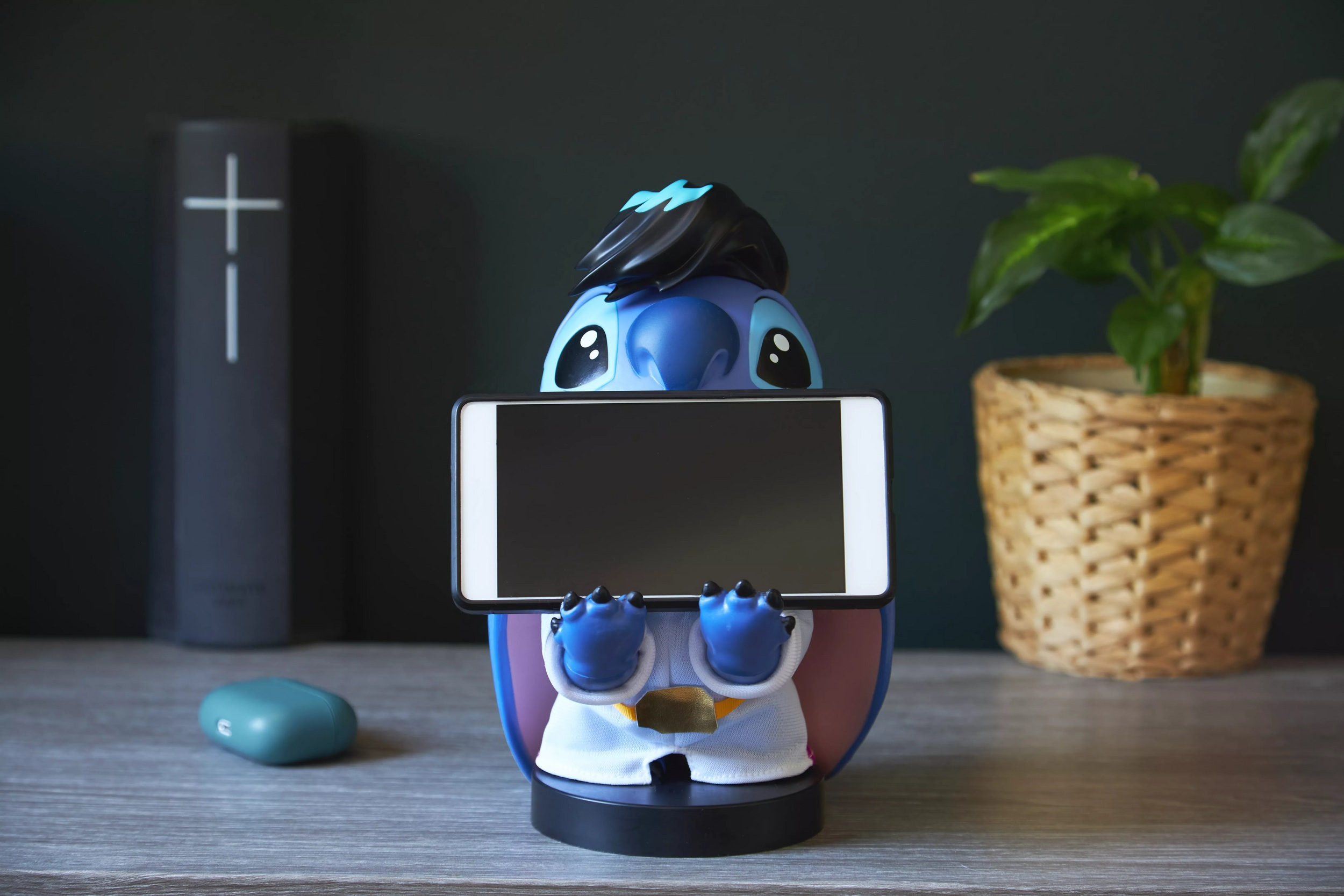 Statuetta di Stitch che tiene un tablet. Su una scrivania di legno con una pianta in vaso e un libro.