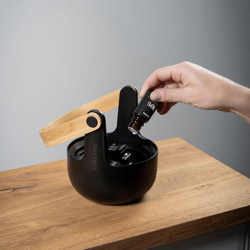 Dłoń wlewa olej z butelki do czarnego dyfuzora. Drewniana powierzchnia.