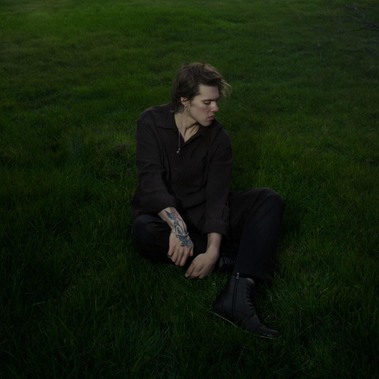 Ein Mann sitzt auf dem Gras und blickt nach unten. Er trägt ein dunkles Hemd und eine Hose. Ein Tattoo ist auf seinem Arm.