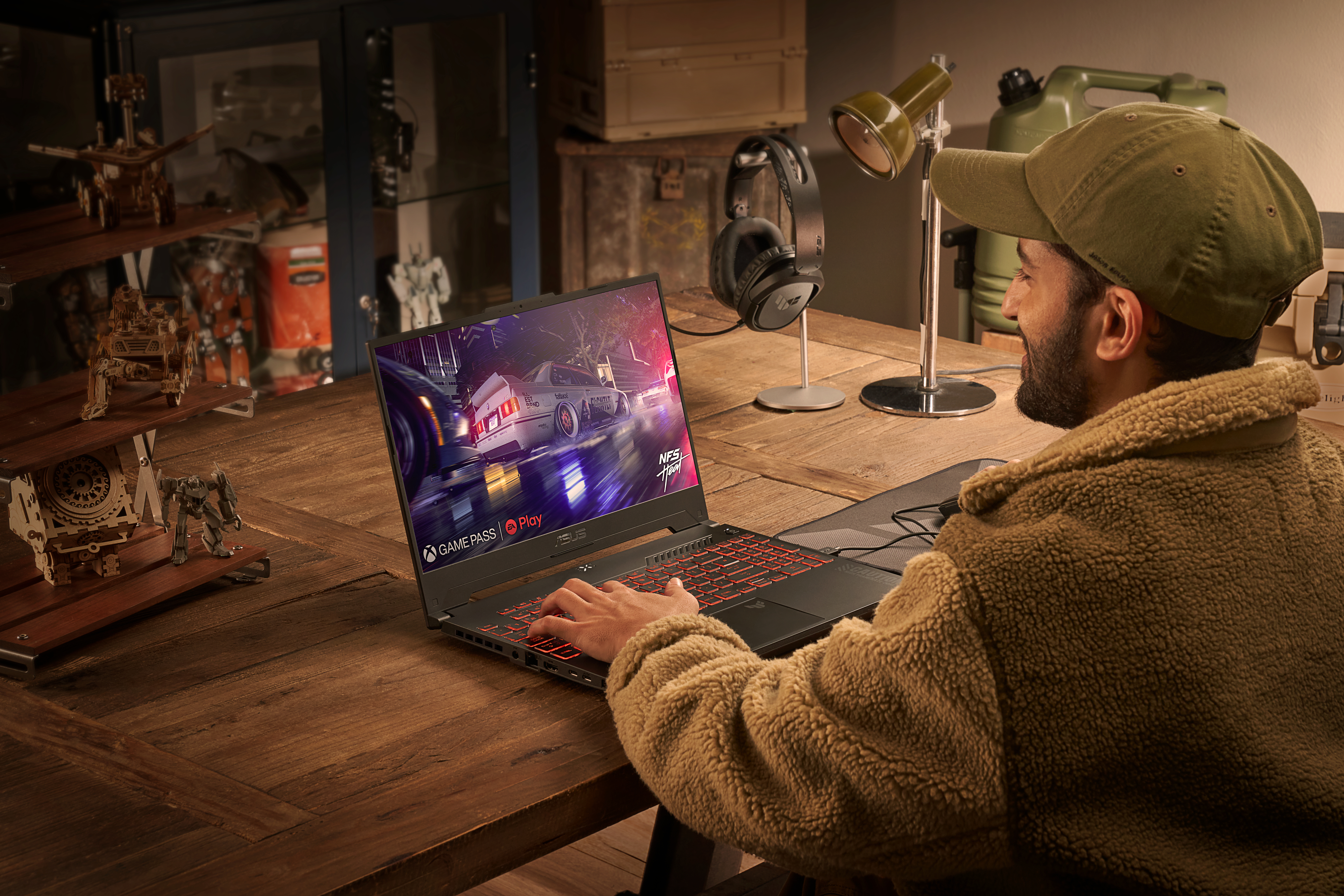 Ein Mann mit Mütze und Jacke spielt auf einem Laptop. Der Laptop zeigt ein Rennspiel. Ein Schreibtisch mit Gegenständen.
