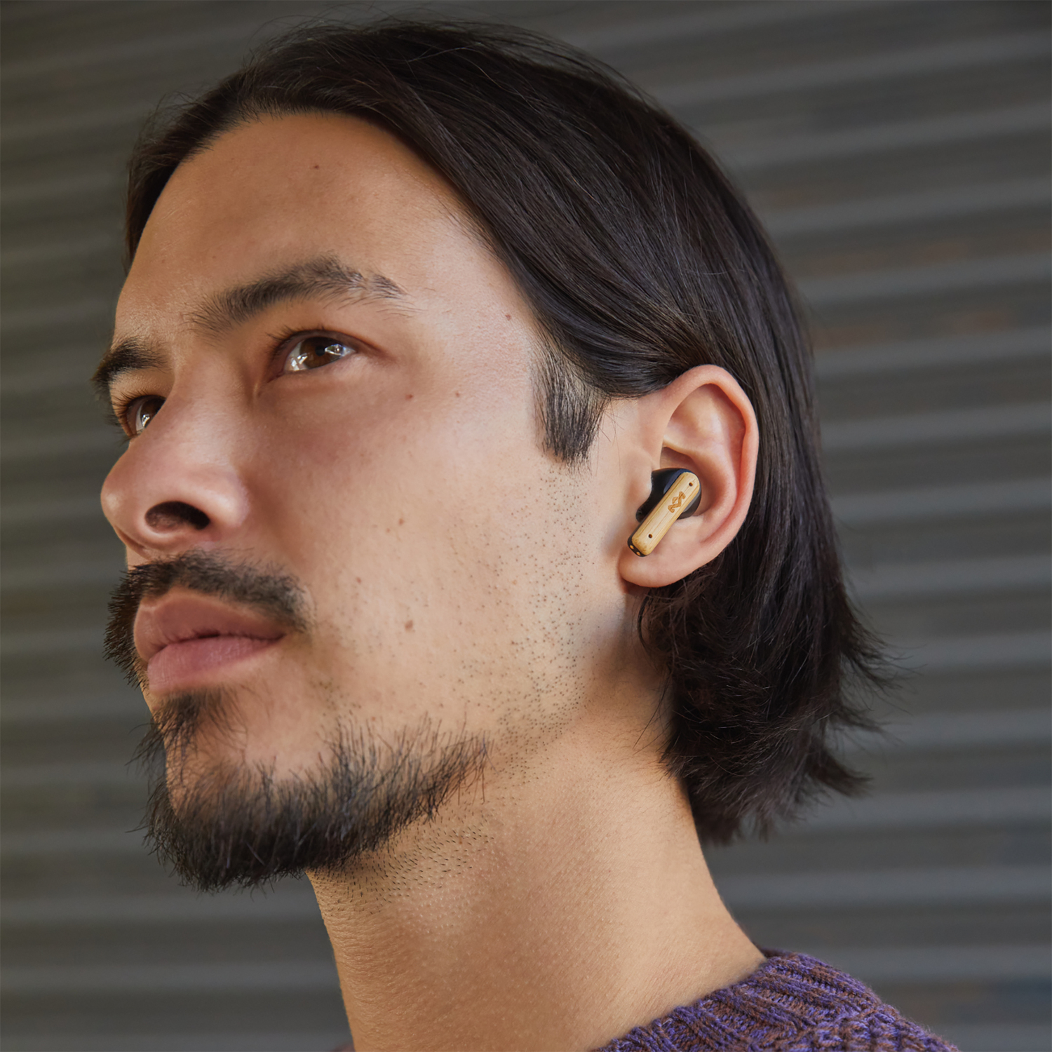 Man wearing purple shirt with an earbud in his ear. Background is a grey wall.