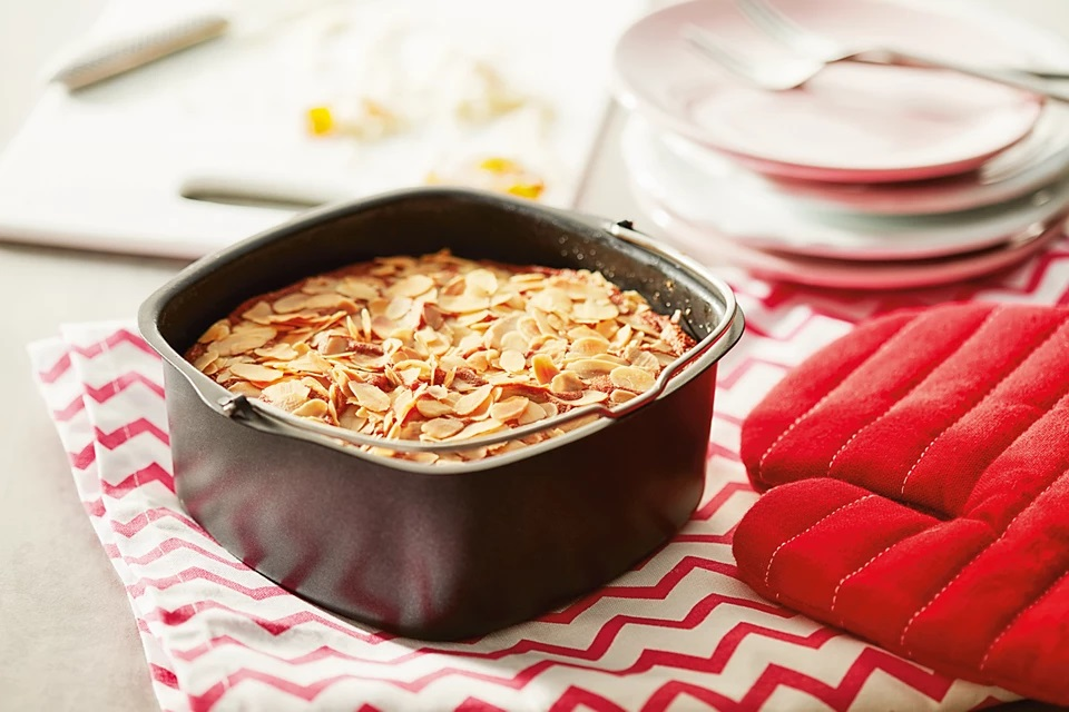 Ciasto w czarnej kwadratowej patelni, posypane płatkami migdałów, ułożone na ściereczce z czerwonymi rękawicami kuchennymi.