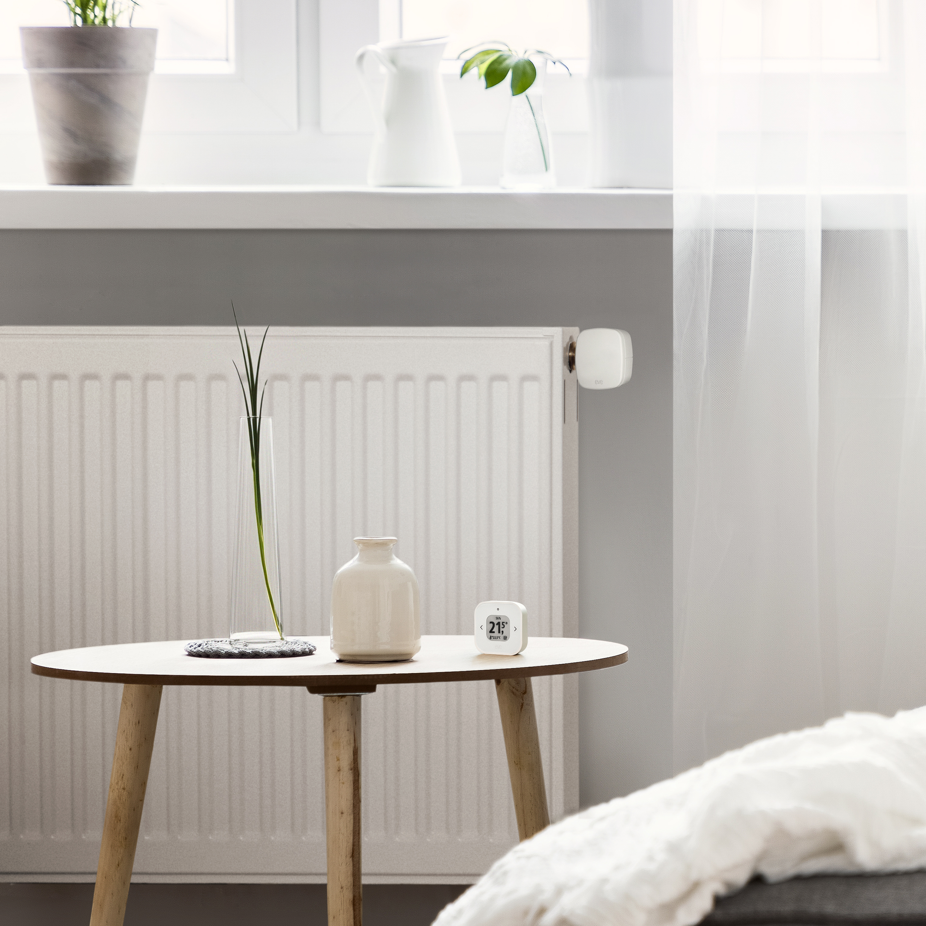 Zimmer mit Heizkörper, Tisch und Smart-Thermostat. Pflanzen am Fenster, weiße Vorhänge.