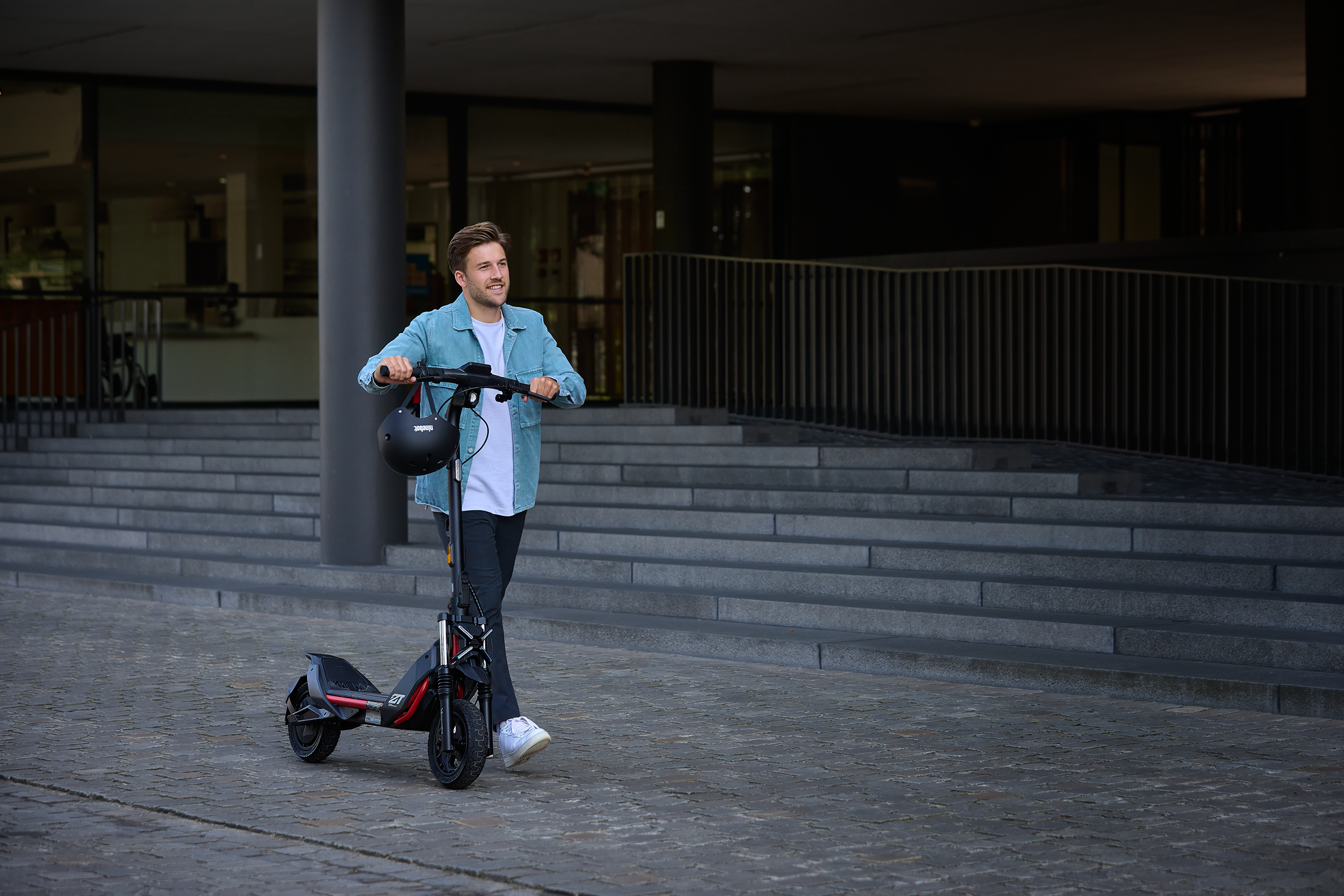 Egy férfi elektromos robogóval sétál egy városi utcán. Sisakot tart.