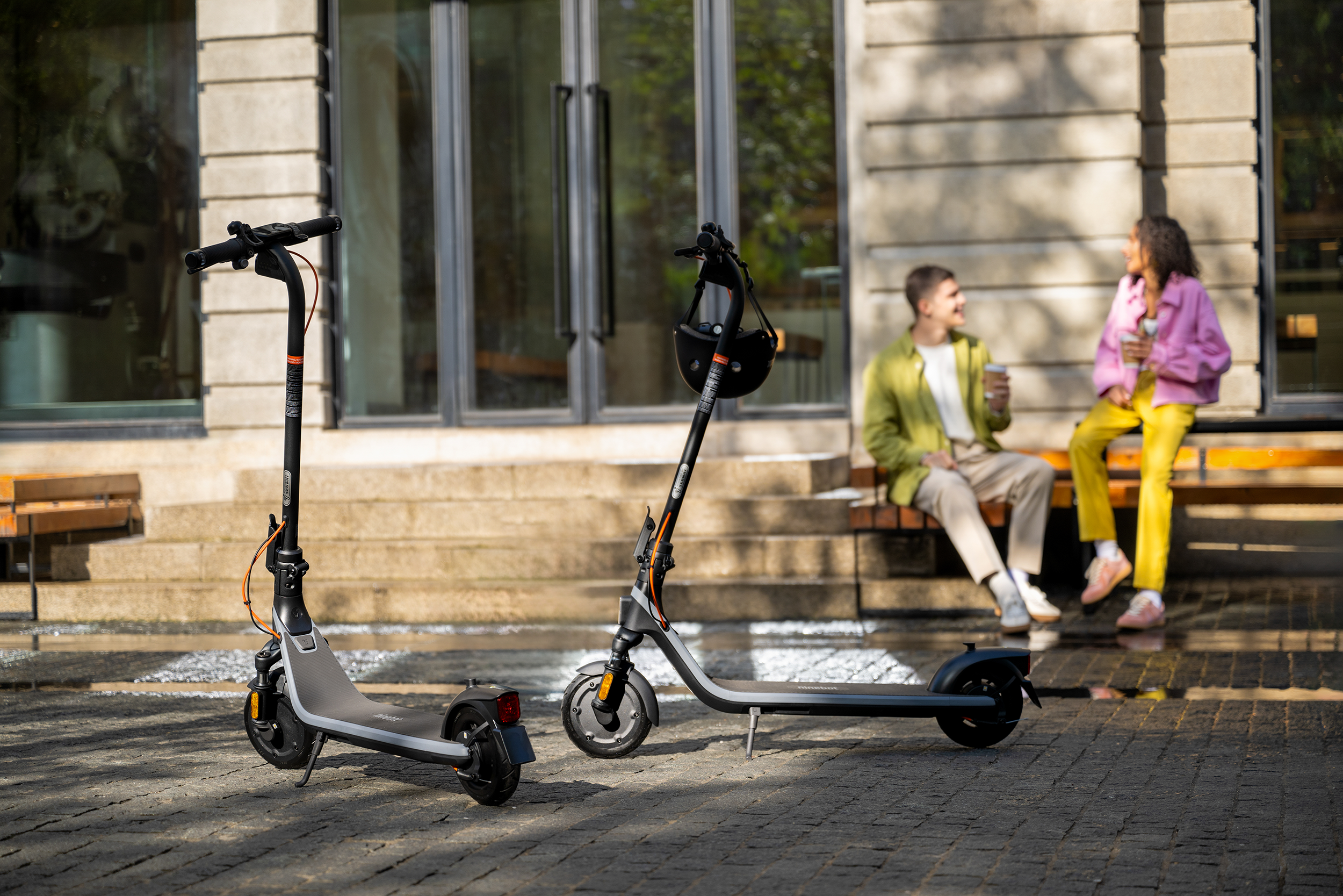 Két elektromos roller parkolva egy burkolt területen, emberek a háttérben.