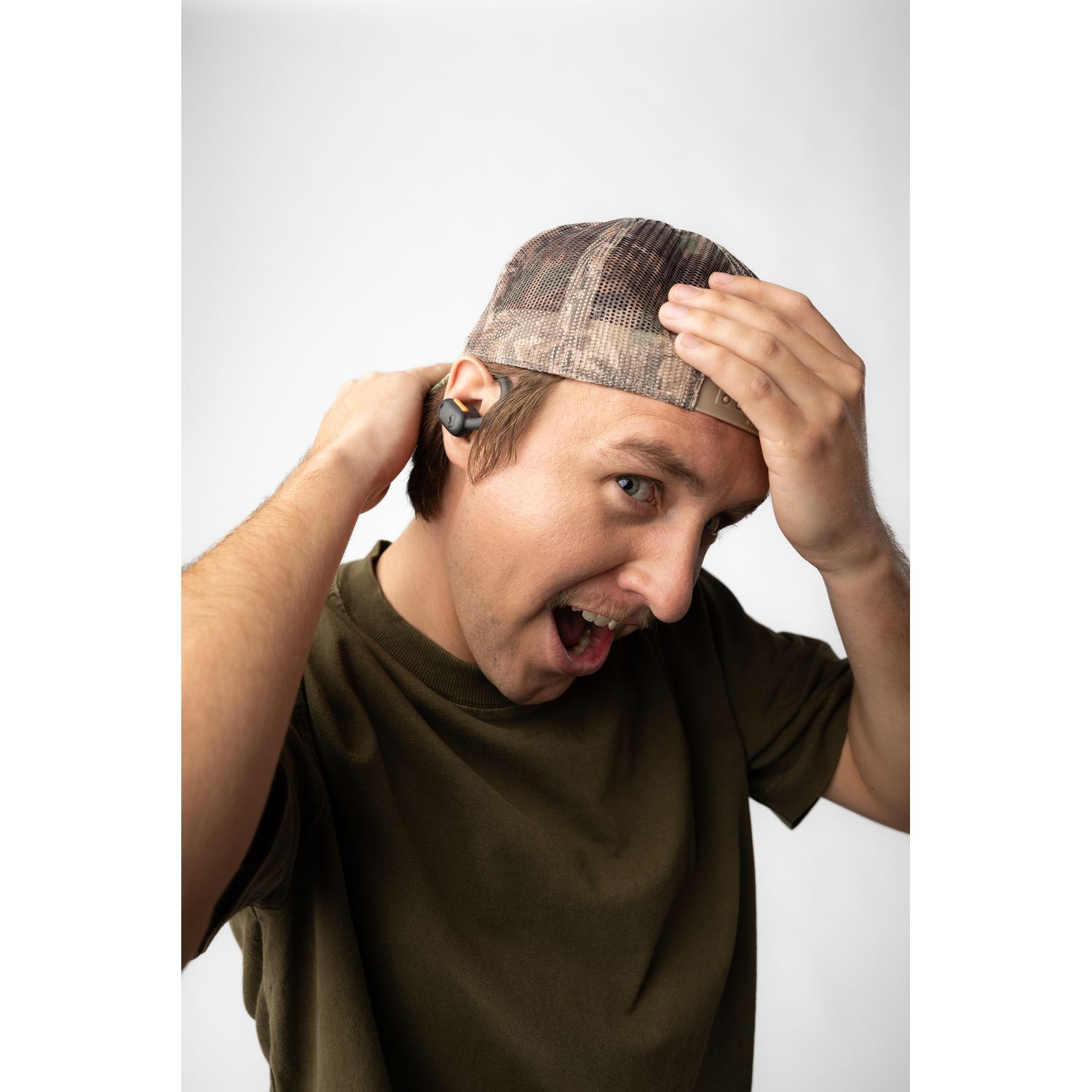 A man wearing a camo hat with earbuds in his ear, looking surprised.