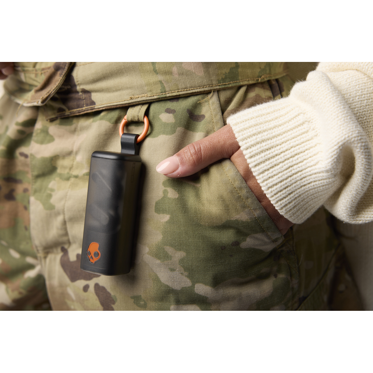Een zwarte Skullcandy-hoes hangt aan een camouflagebroek. Een hand rust in een zak, een crèmekleurige mouw is zichtbaar.