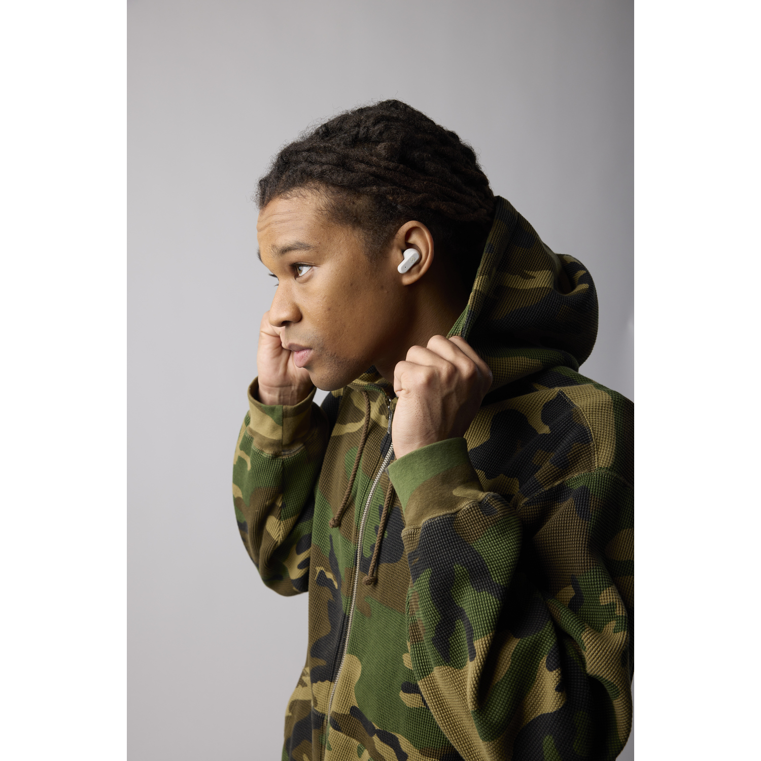Een jonge man met een camouflage hoodie doet witte oortelefoons in, kijkend naar links tegen een grijze achtergrond.