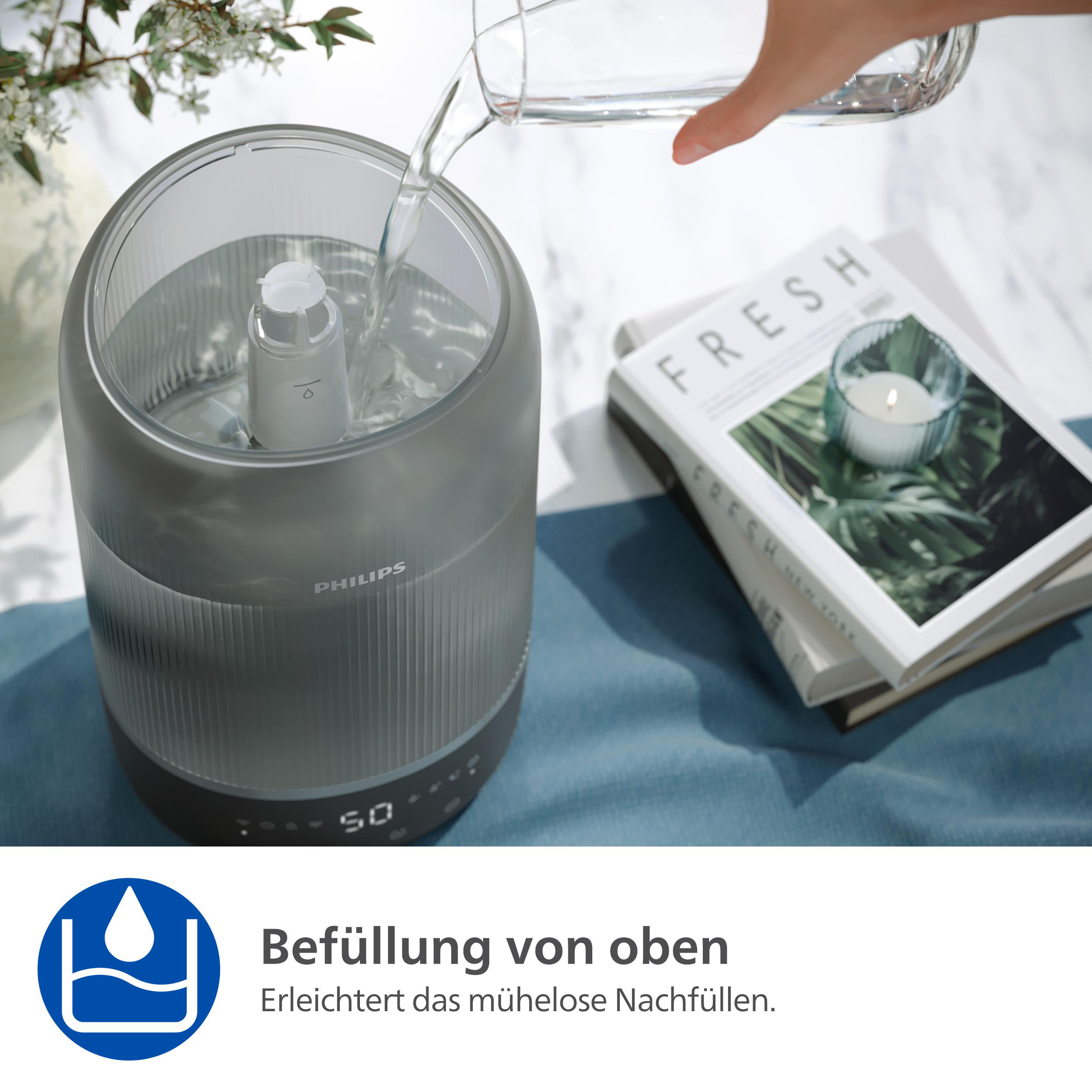Eine Hand gießt Wasser in einen grauen Luftbefeuchter. Ein Buch und Blumen liegen auf dem Tisch.