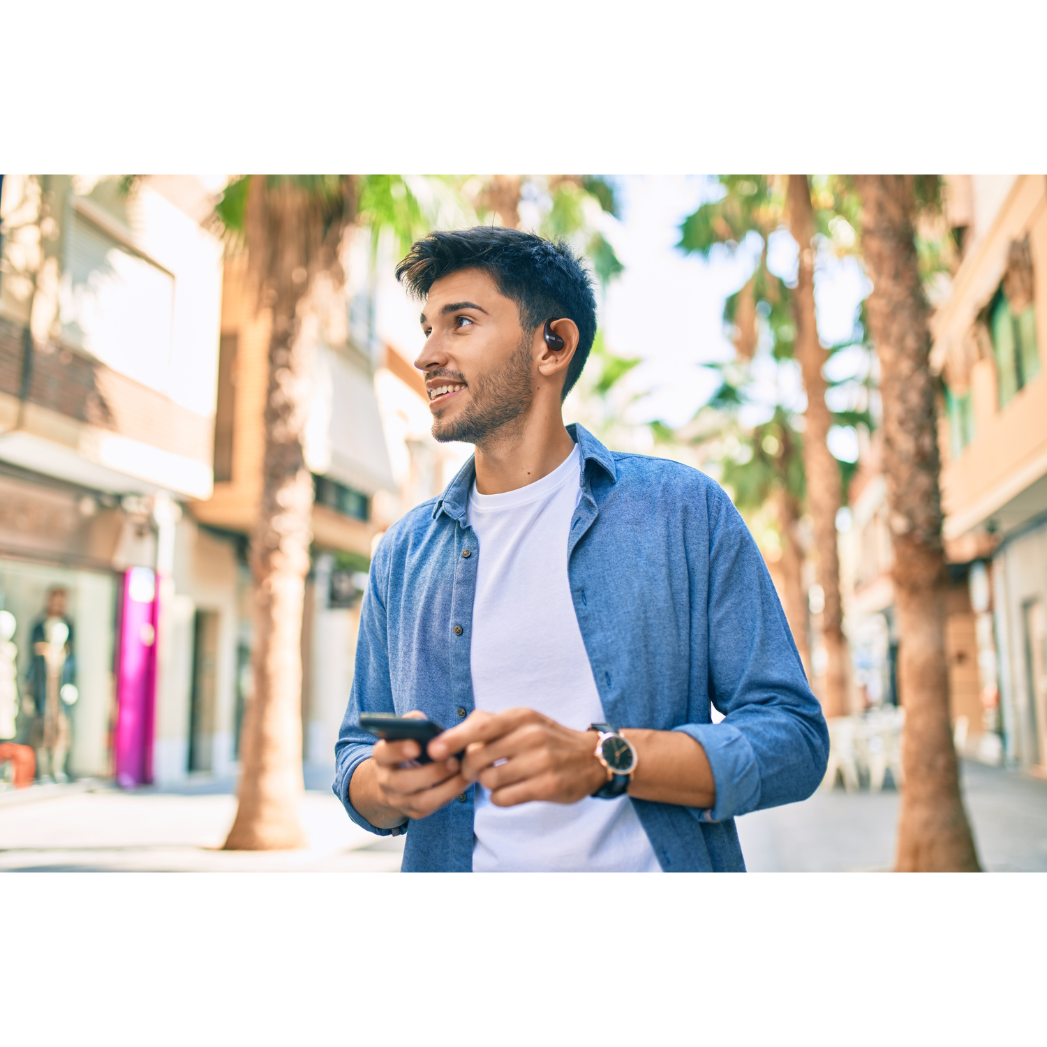 Homme en chemise bleue marchant dans une rue de la ville, portant des écouteurs et tenant un téléphone.
