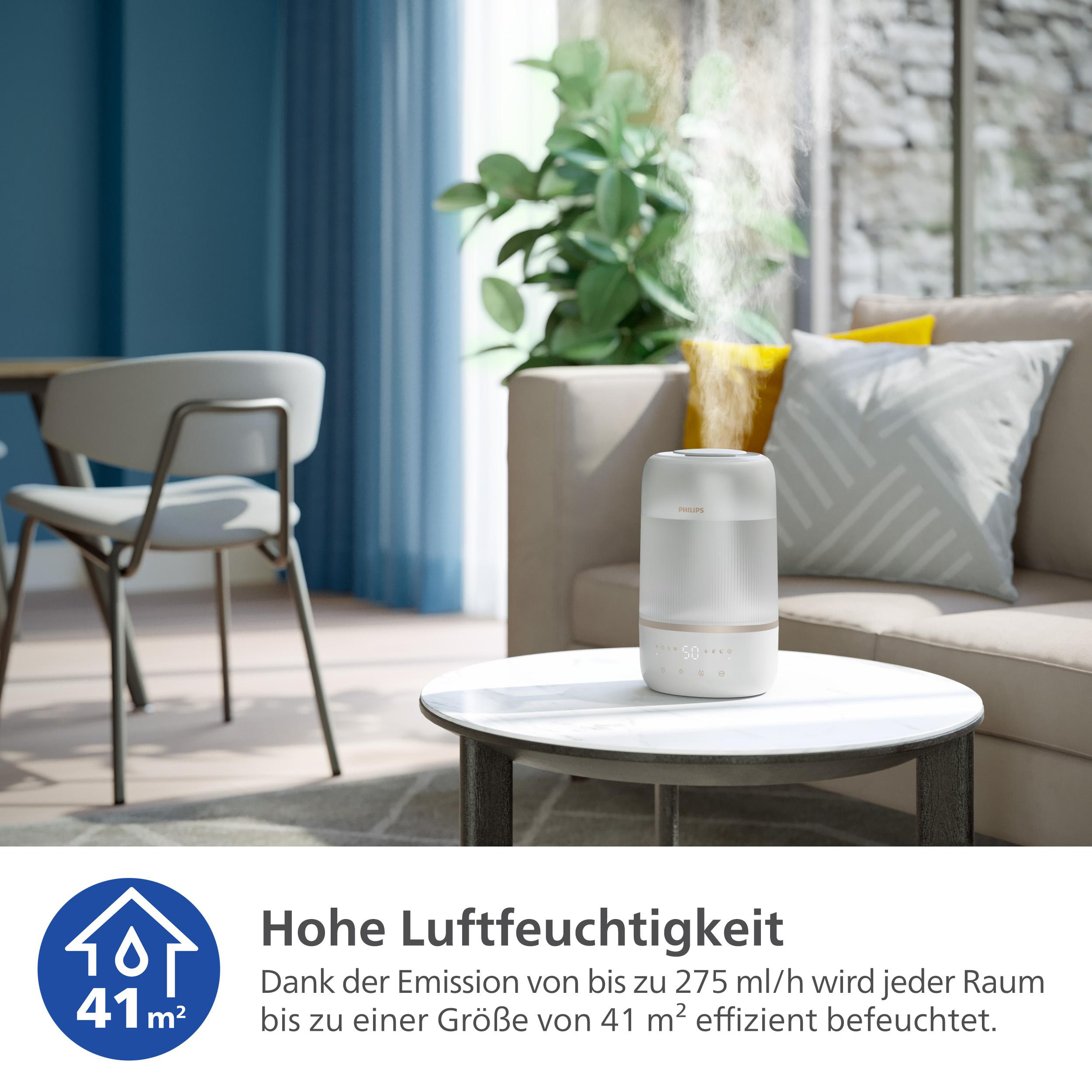 Ein weißer Luftbefeuchter auf einem Tisch, mit aufsteigendem Dampf. Eine blaue Wand und eine Person sind sichtbar.