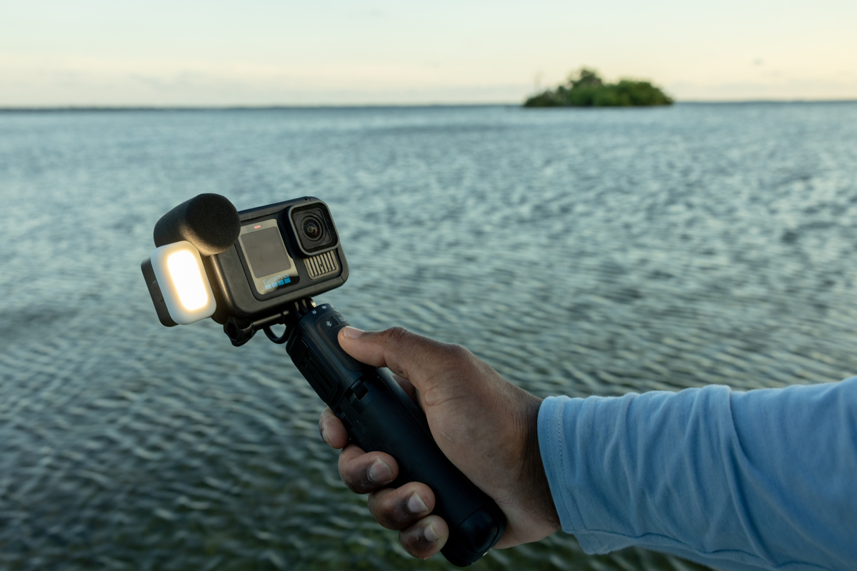 Eine Person hält eine schwarze Kamera mit einem Lichtaufsatz über Wasser. Eine Insel ist in der Ferne.
