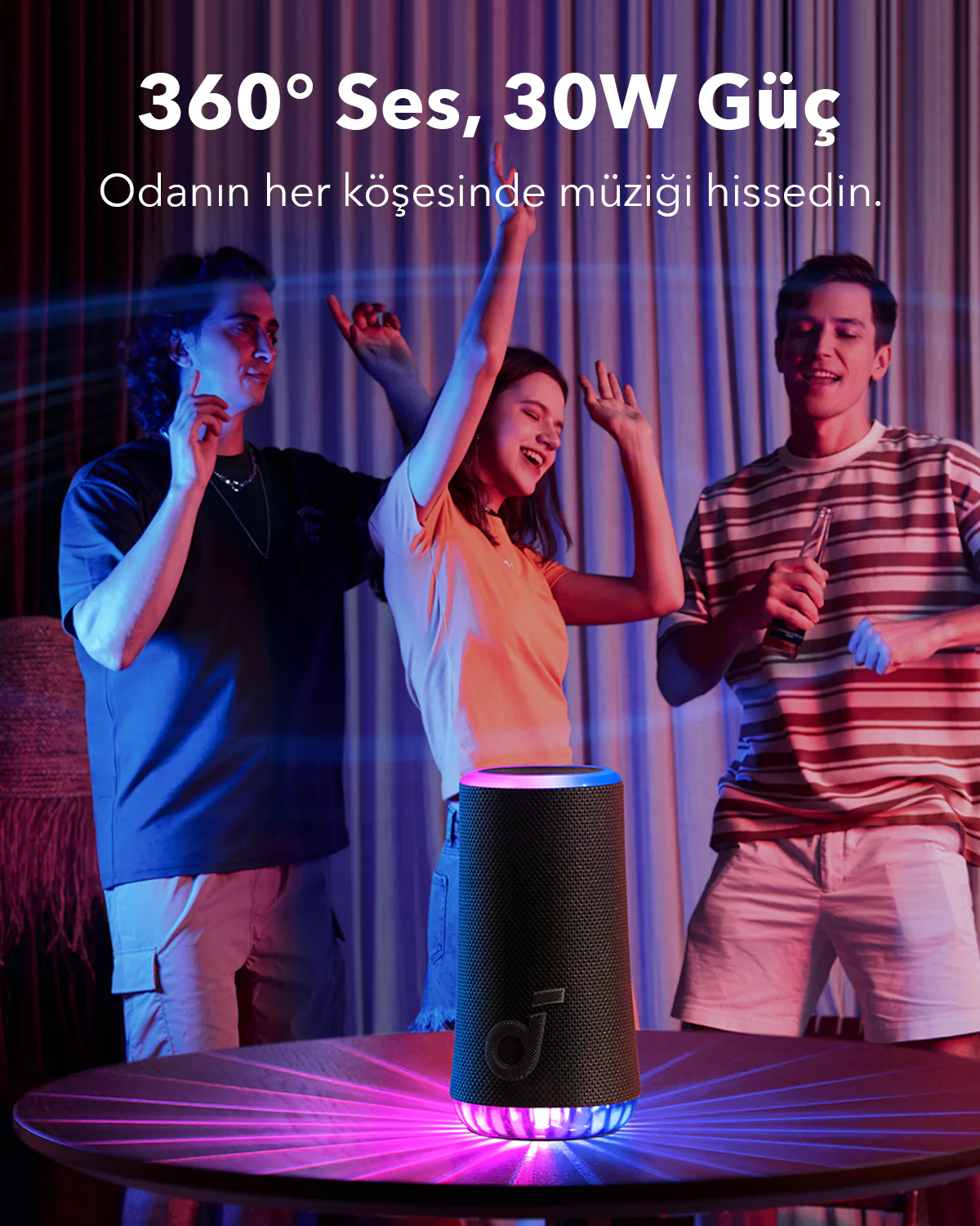 Üç kişi bir masadaki hoparlörün yakınında dans ediyor. Hoparlörün renkli ışıkları var. Arka plan bulanık.