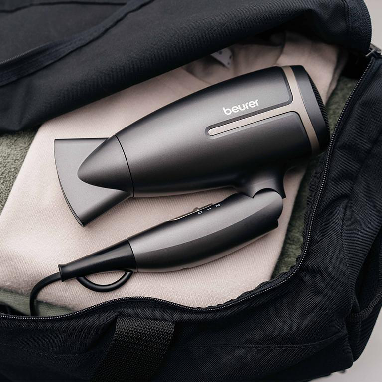 Ein schwarzer Haartrockner liegt in einer schwarzen Tasche auf gefalteter Kleidung. Der Trockner ist grau und schwarz.