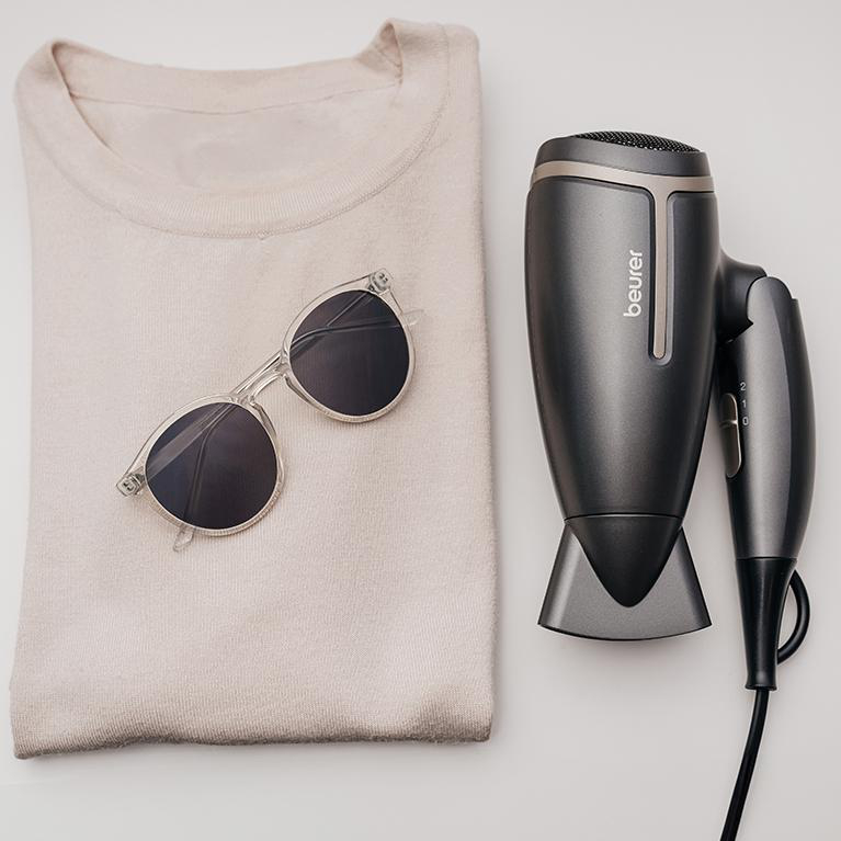 Sonnenbrille und ein Haartrockner auf einem gefalteten Pullover. Weiße Oberfläche. Neutrale Farben.