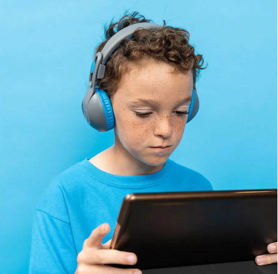 Chłopiec w słuchawkach patrzy na tablet. Ma piegi i niebieską koszulę na niebieskim tle.