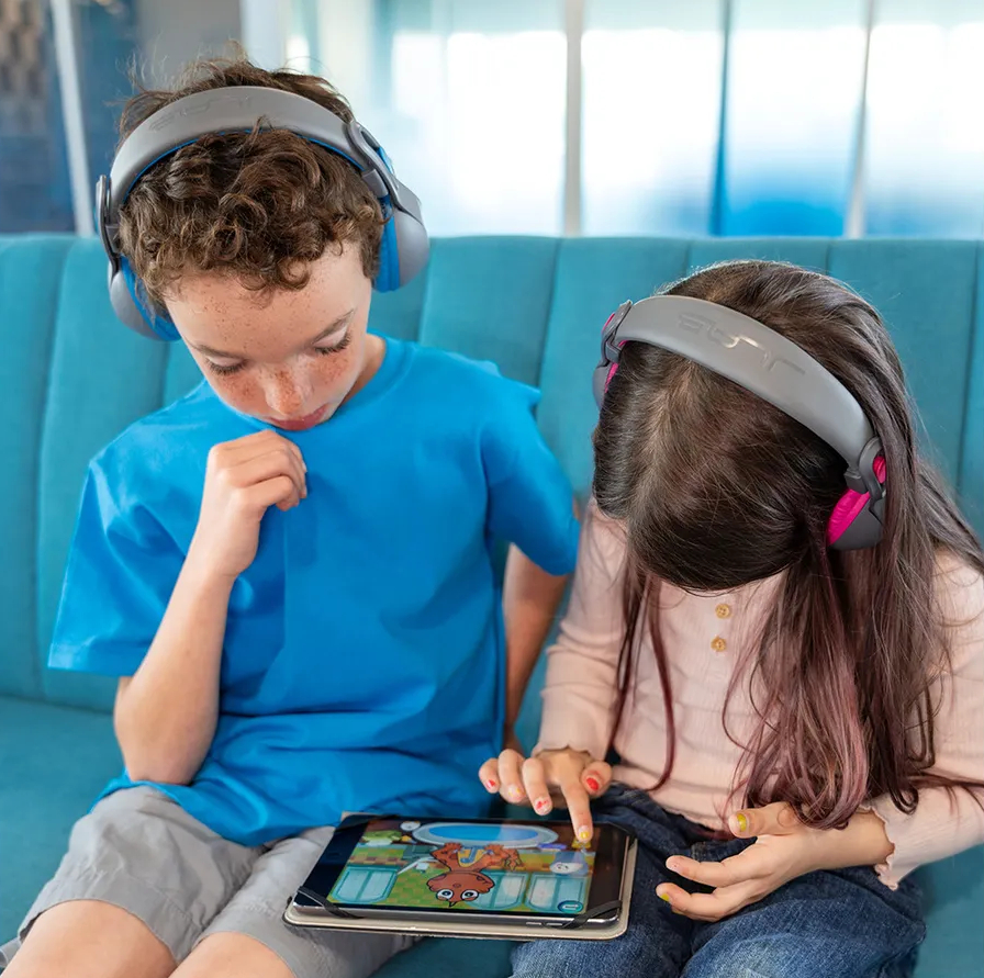 Dwoje dzieci siedzi i patrzy na tablet. Mają na sobie słuchawki.