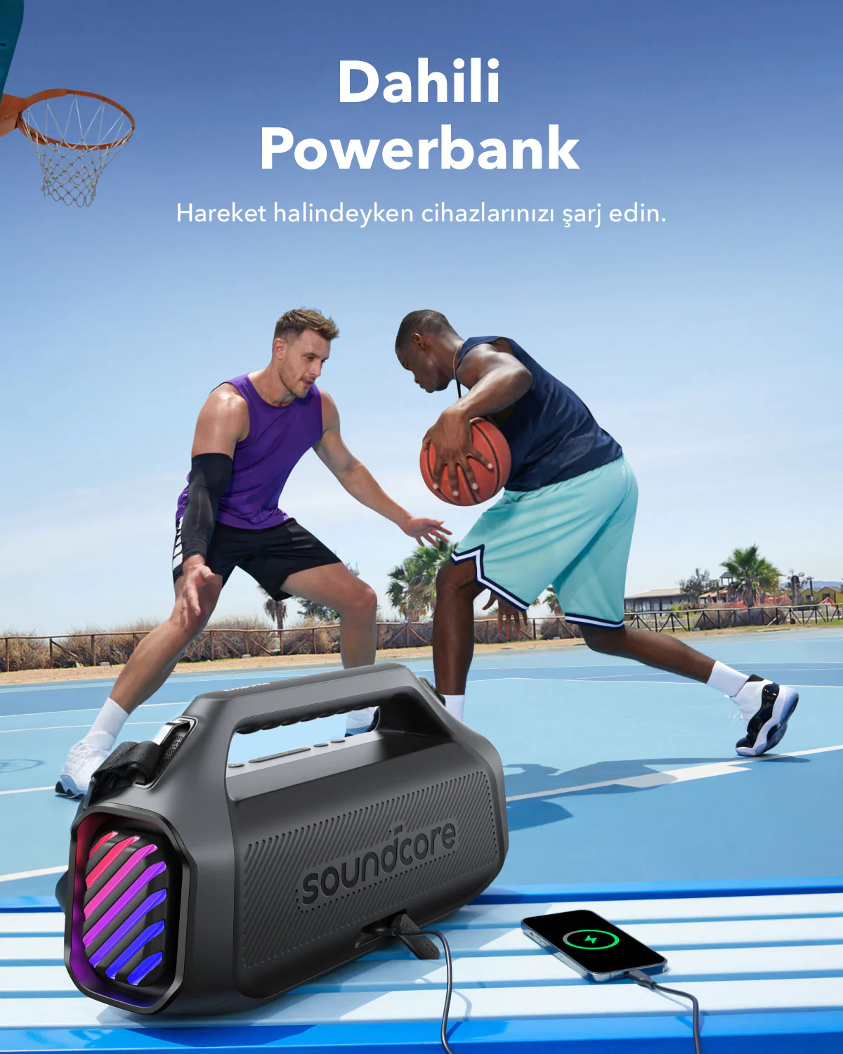 İki adam, bir Soundcore powerbank'inin yanında basketbol oynuyor. Bir telefon şarj oluyor. Arka planda palmiye ağaçları olan basketbol sahası.