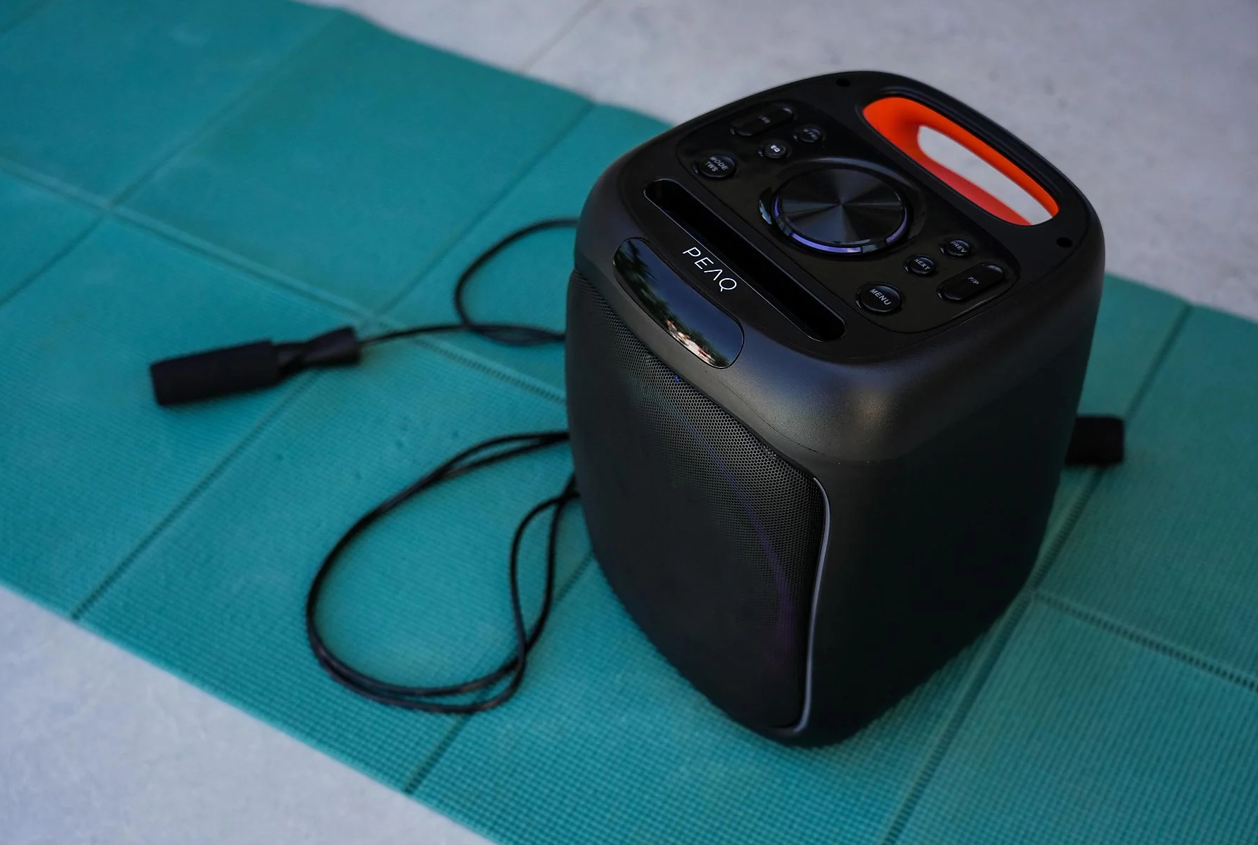 Czarny głośnik z pomarańczowym uchwytem na zielonej macie. Obok skakanka.