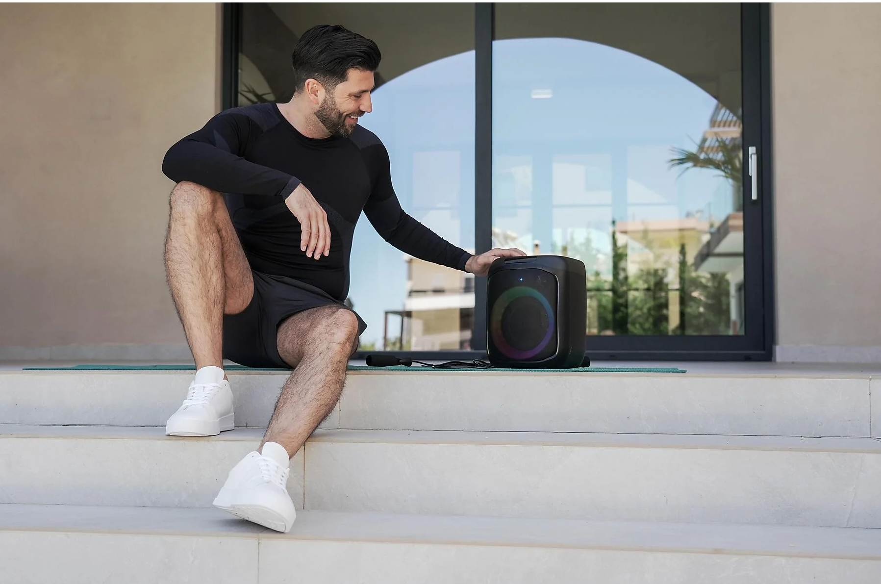 Mężczyzna siedzi obok czarnego głośnika. Uśmiecha się i ma na sobie strój sportowy.