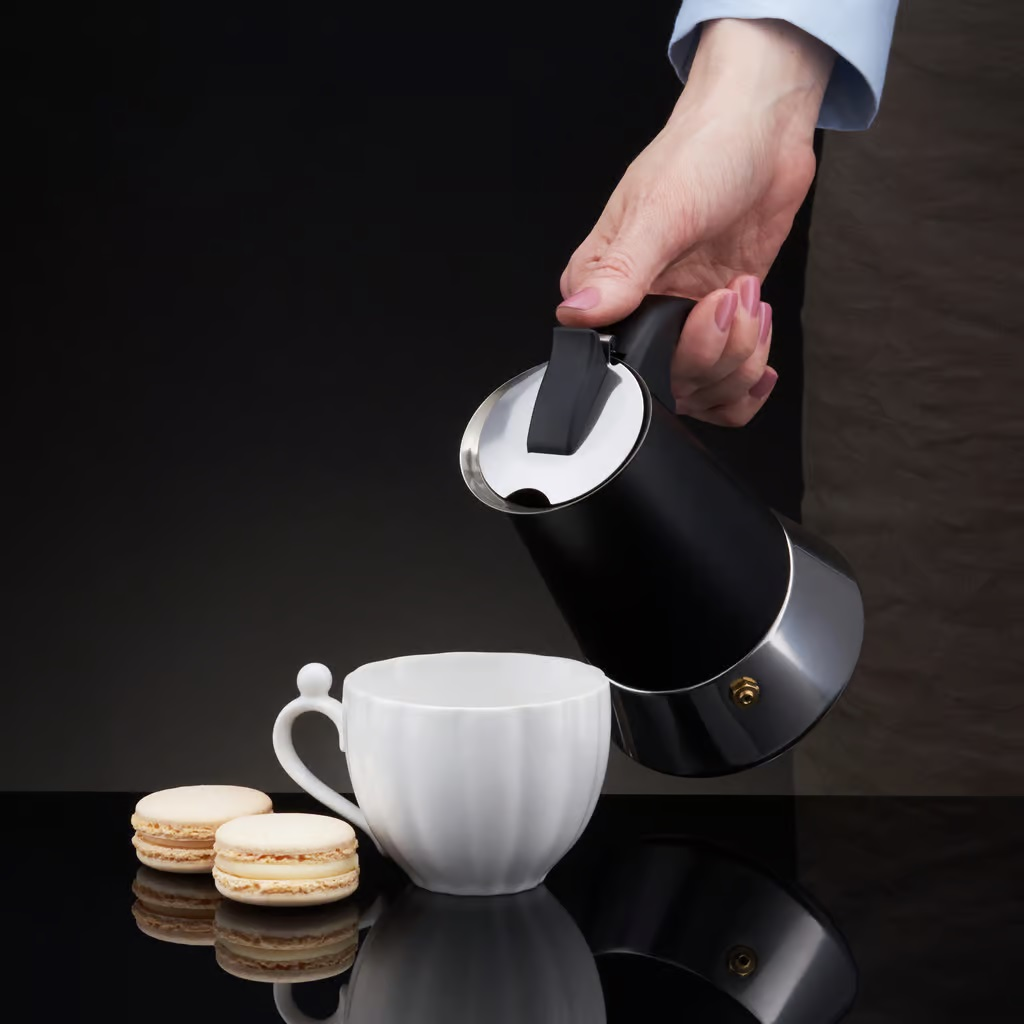 Ręka nalewa kawę z czarno-srebrnego dzbanka do białego kubka. Makaroniki obok.