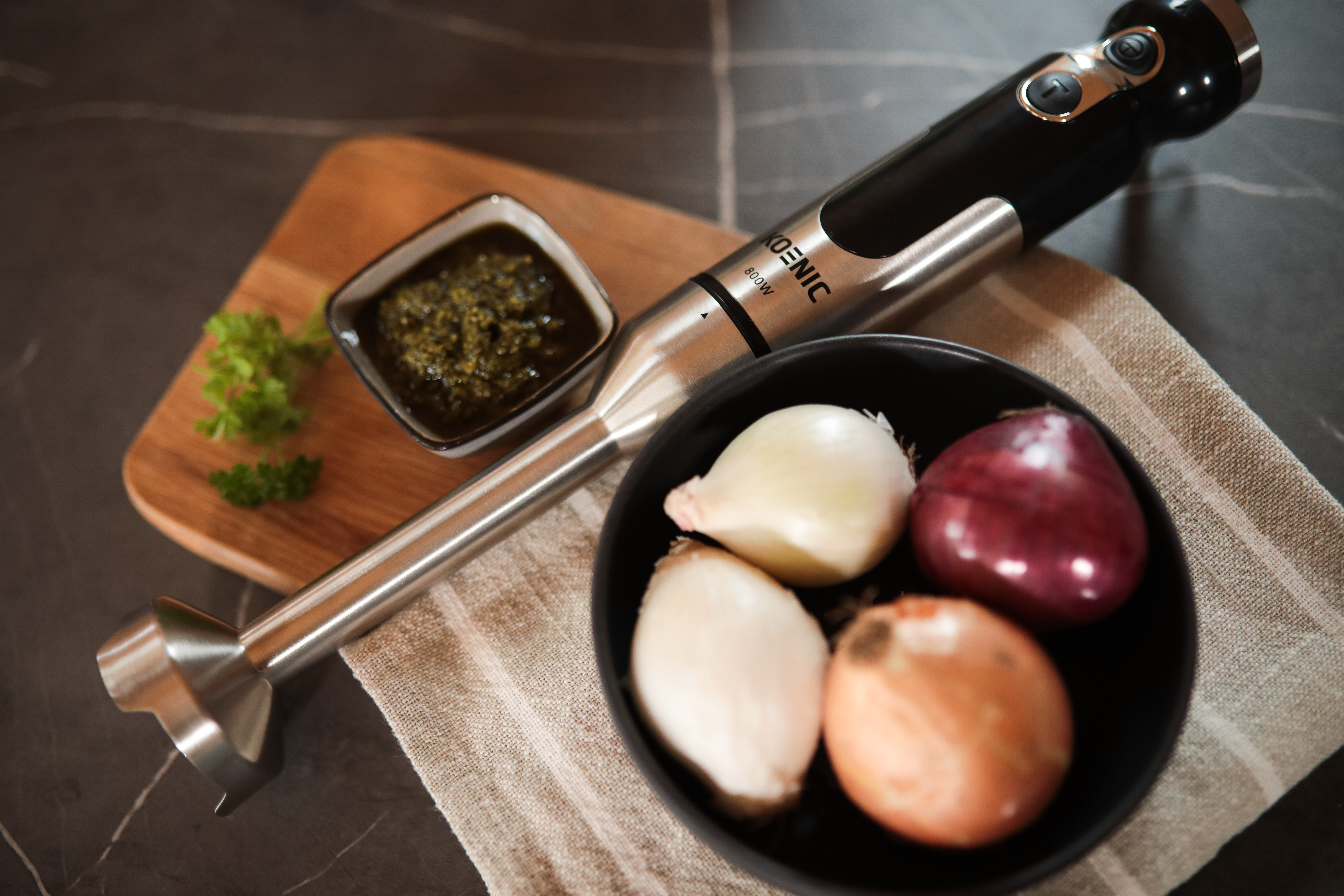 Batidora de mano con ingredientes: cebollas, pesto, perejil, en tabla y bol negro.
